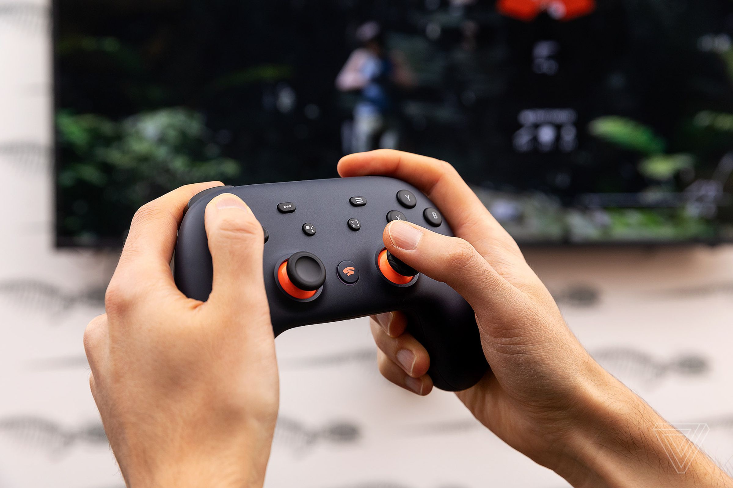 Two hands hold a Stadia controller in front of a TV.
