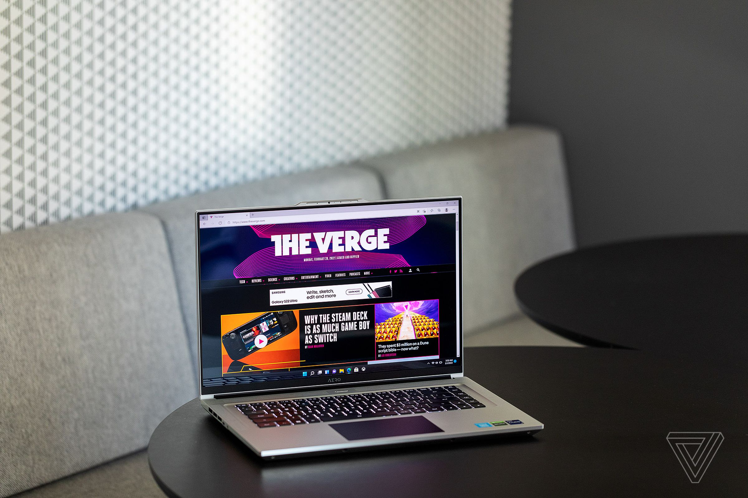 The Gigabyte Aero 16 angled to the right on a cafe table. The screen displays The Verge homepage.