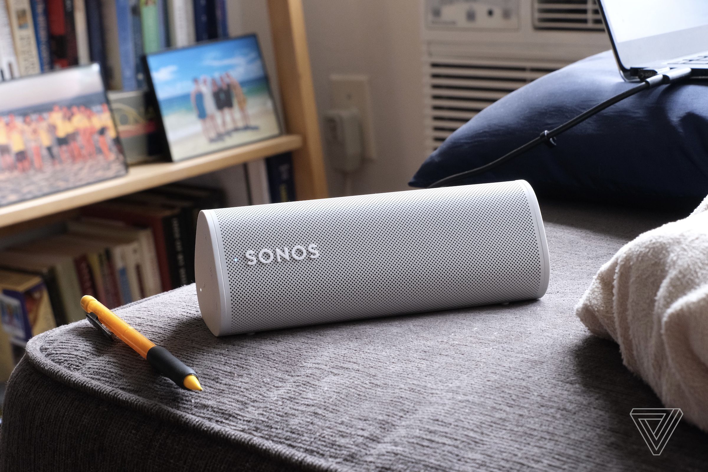 The white Sonos Roam sitting on a bed with picture frames behind it.