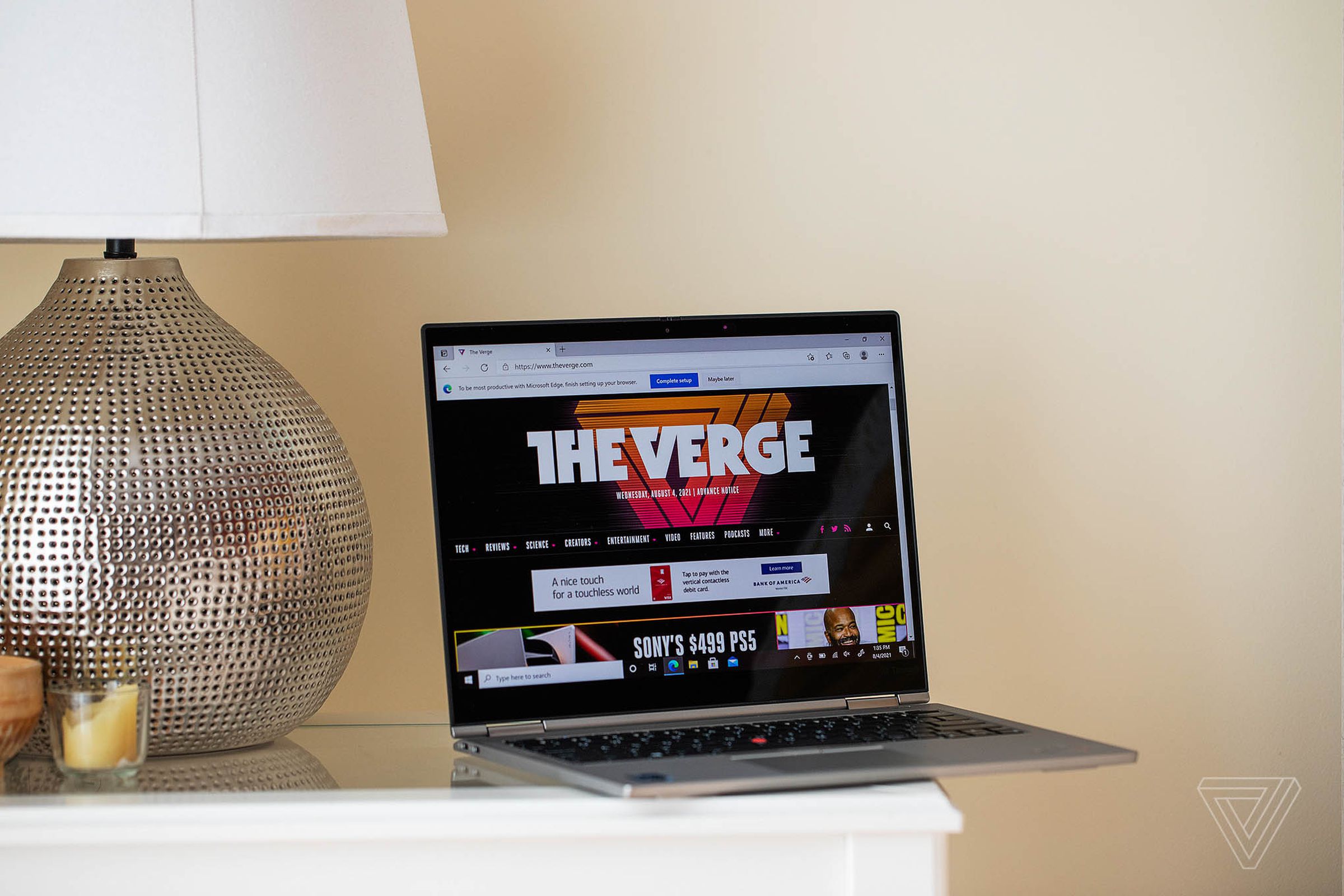 The Lenovo ThinkPad X1 Titanium Yoga open on a bedside table next to a lamp, angled to the right. The screen displays The Verge’s homepage.