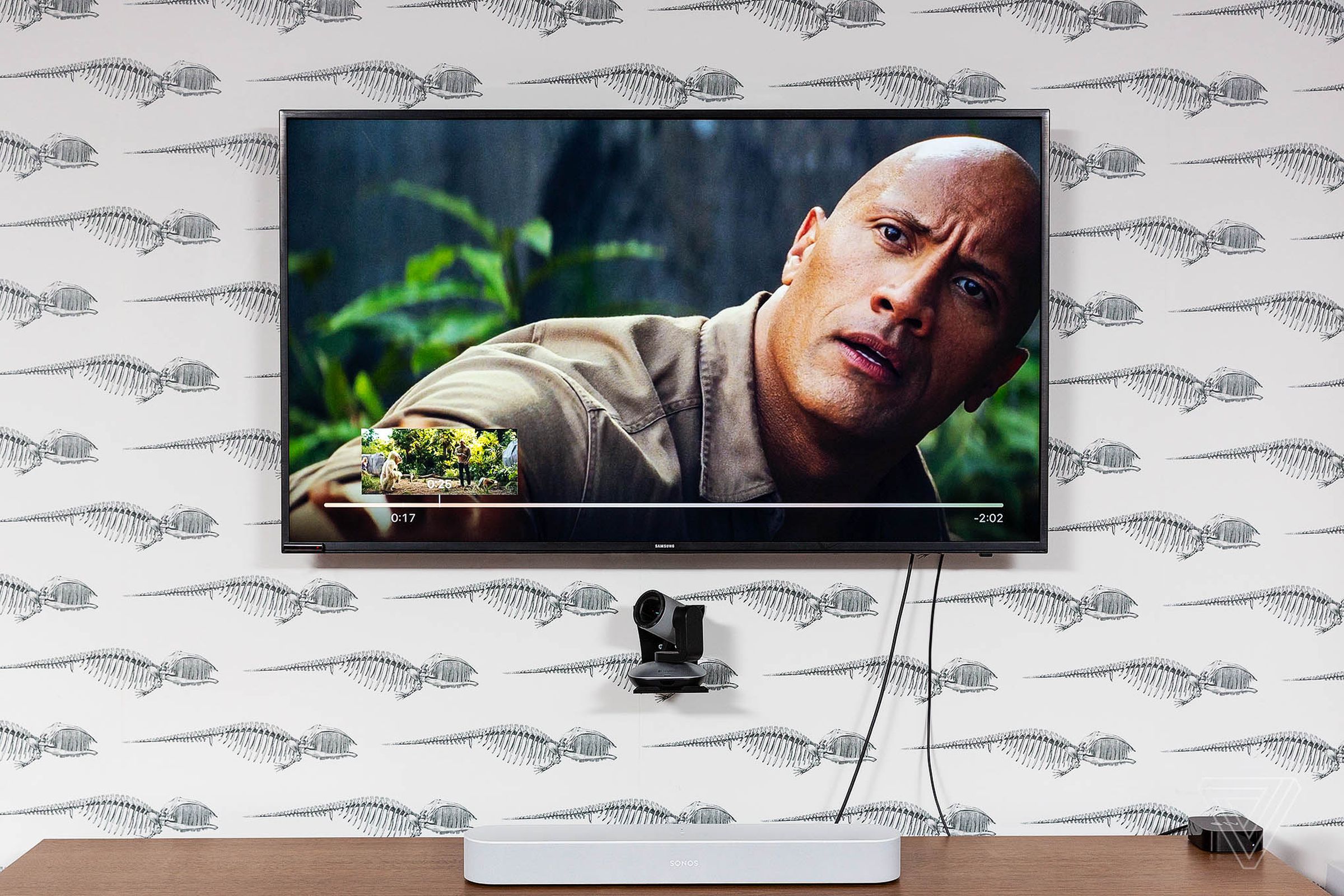 The Sonos Beam shown on an entertainment center with Dwayne “The Rock” Johnson displayed on a TV.