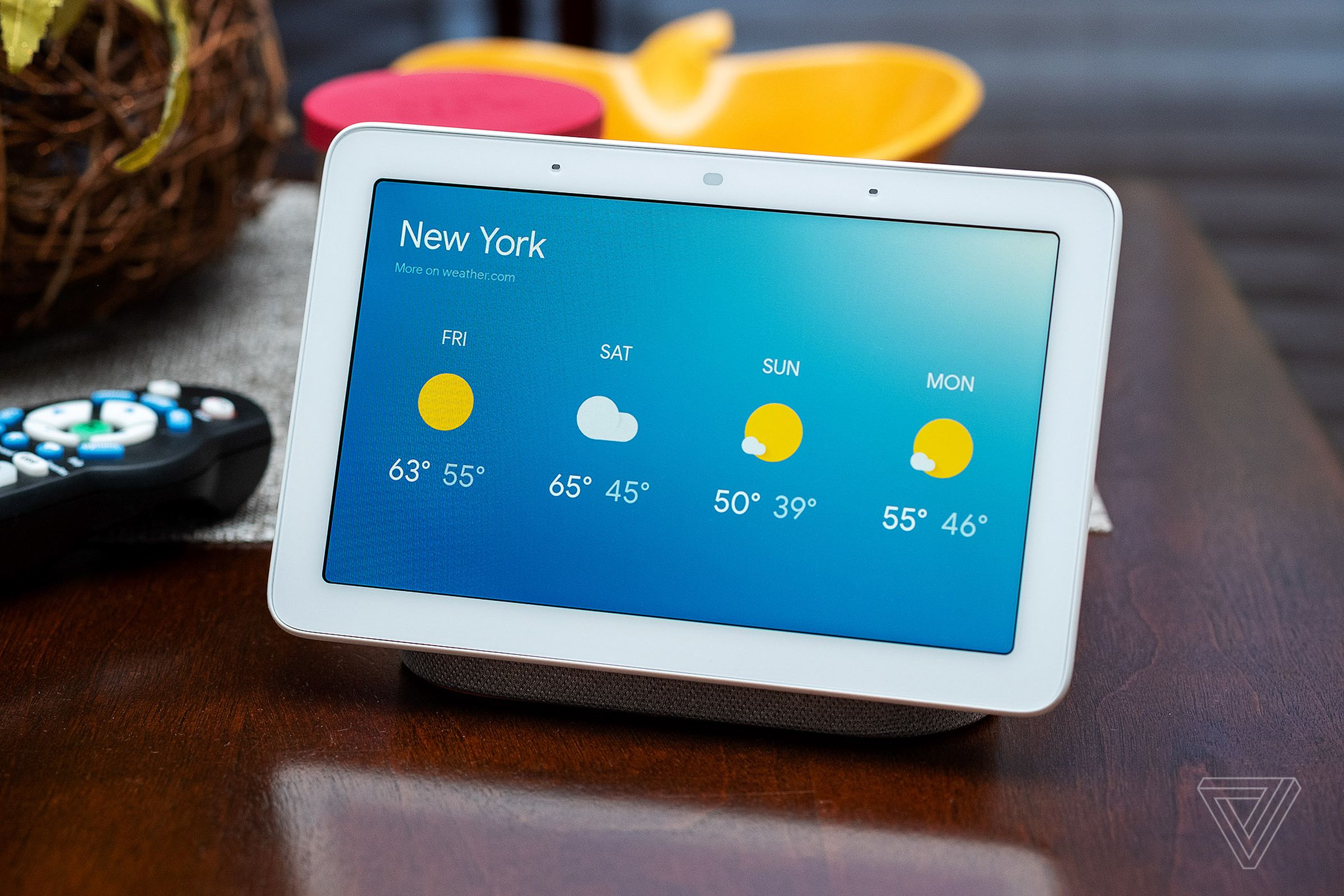 A first-gen Google Nest Hub showing the weather, sitting on a desk.