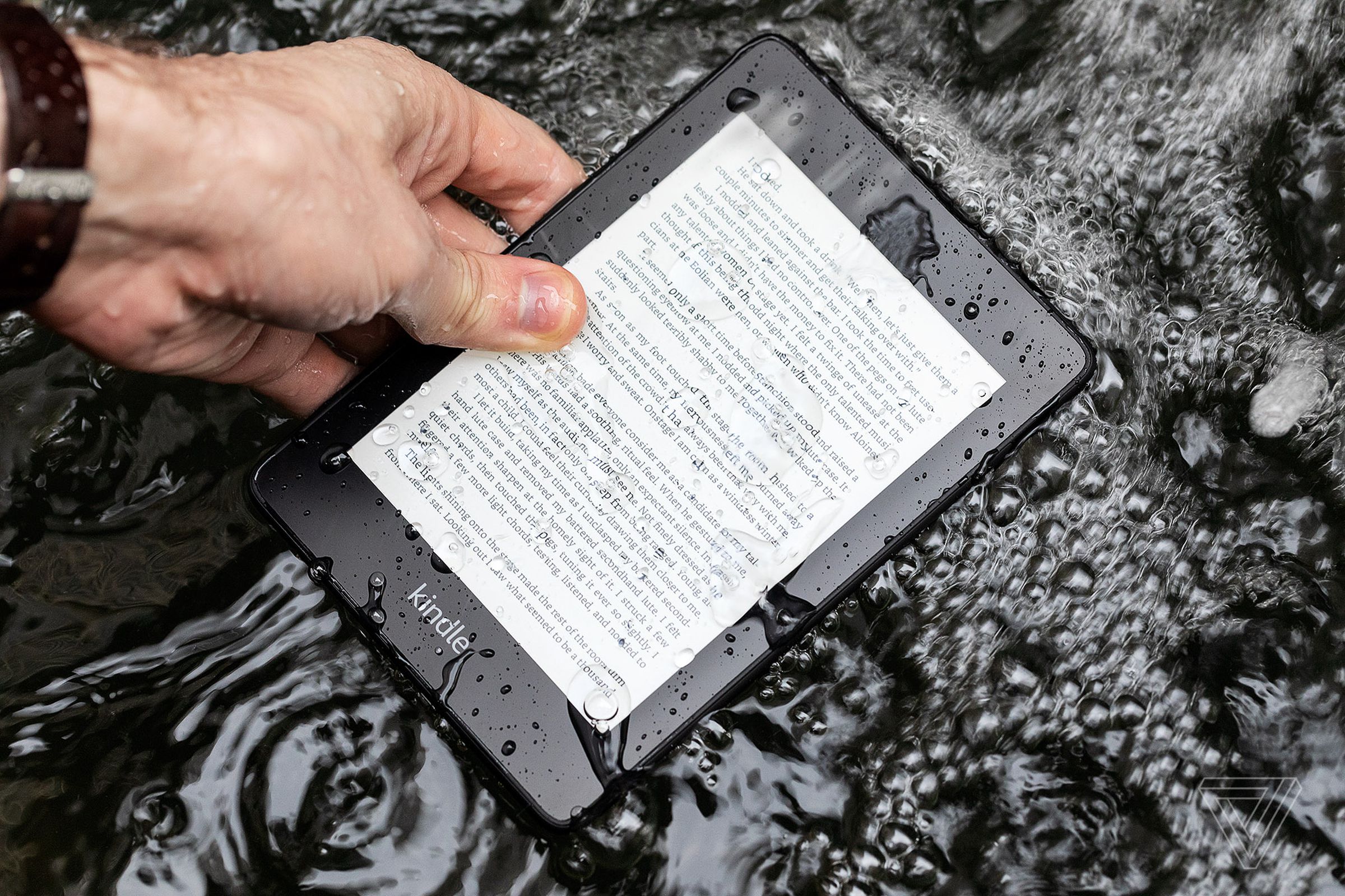 A hand holding a very wet Kindle Paperwhite as it lies on the ground.
