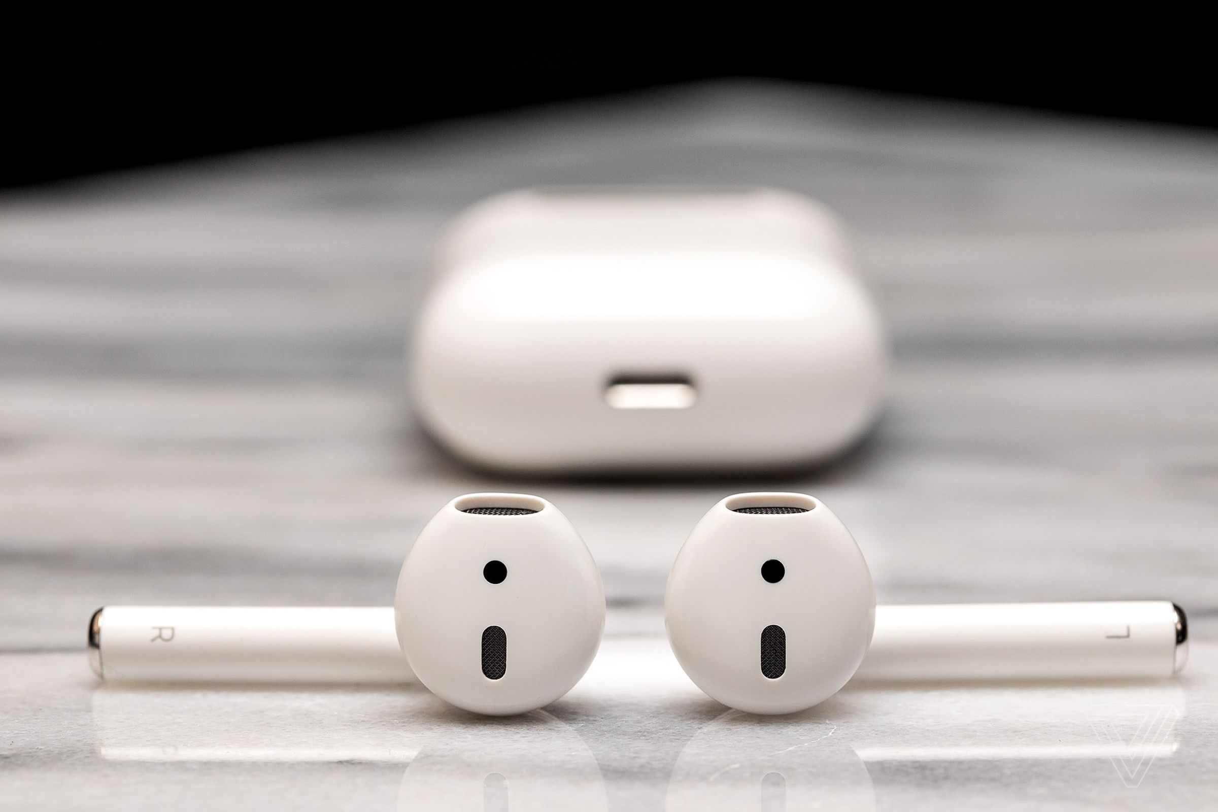 A close-up pictures of a set of second-gen Apple AirPods on a marble tabletop. Their charging case is set behind them, blurred out of focus, with its bottom Lightning port visible.