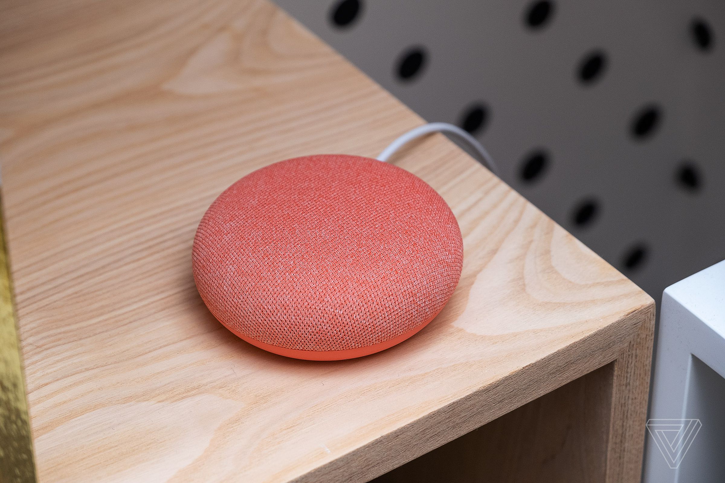 A coral colored Google Nest Mini smart speaker sitting on a wood desk.