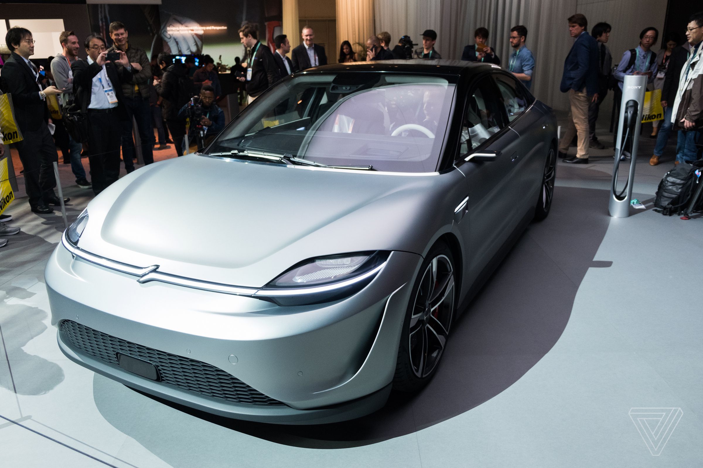 Sony’s Vision-S car is on the CES show floor with lots of people surrounding it. The car, a four-door midsize sedan, is silver with a black top and all-glass roof.