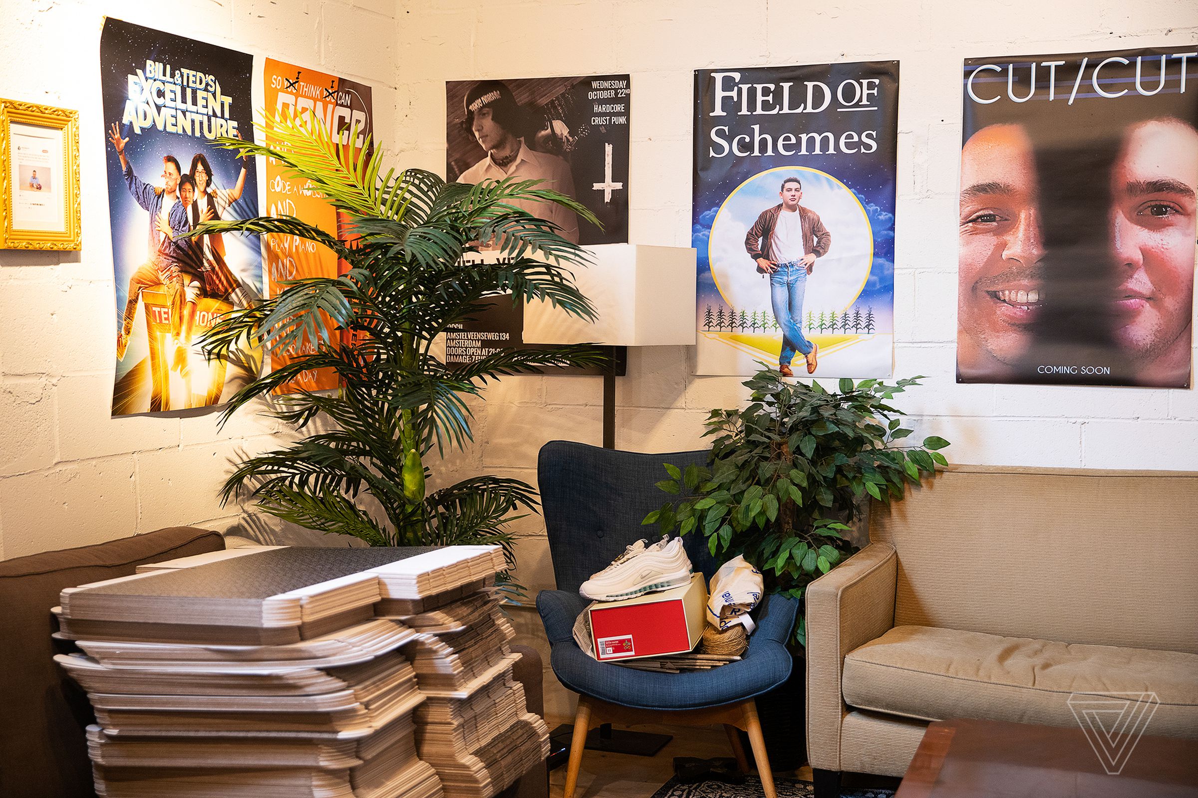 Couches and chairs are in crowded corner of a room with posters on the walls, a pair of sneakers and other things in a pile.
