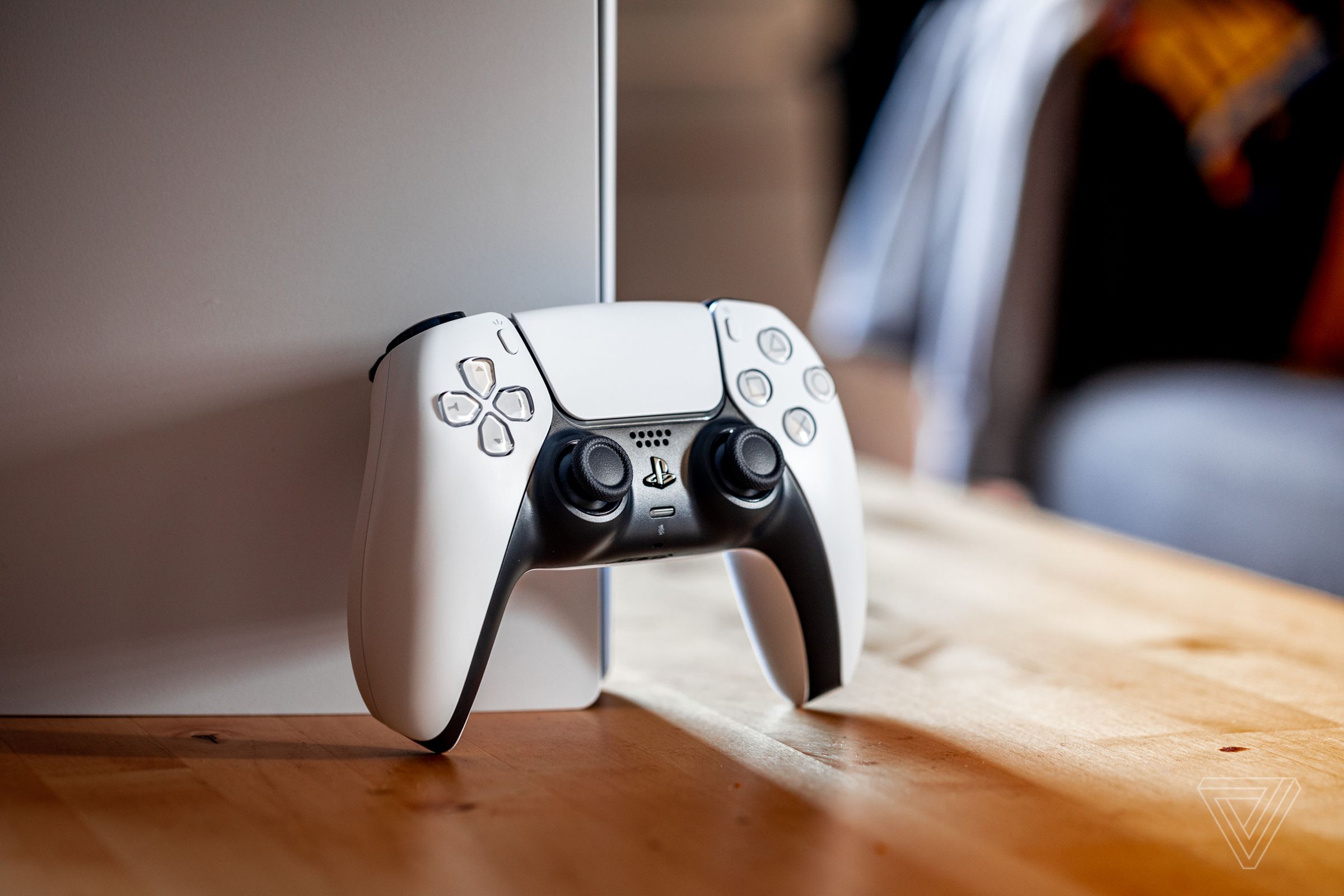 A white Sony DualSense controller resting against a PlayStation 5 console on a wood table.