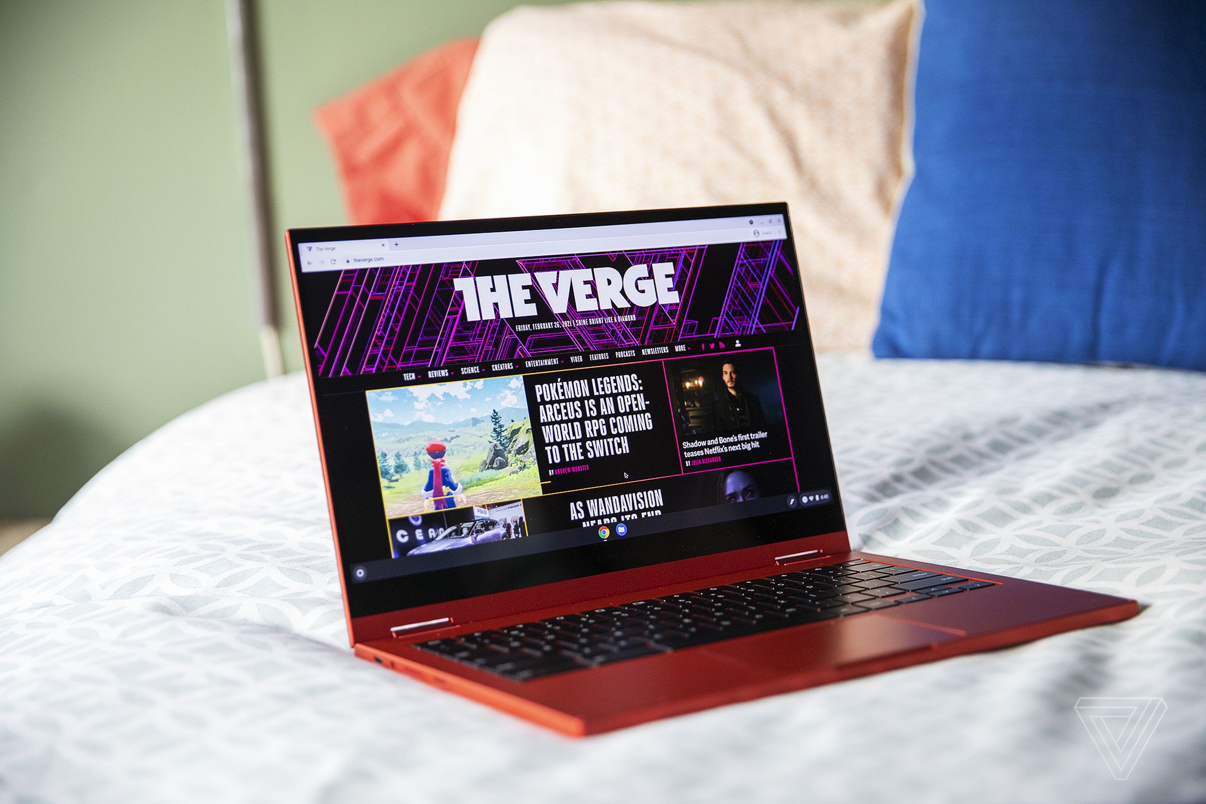 The Samsung Galaxy Chromebook open, angled slightly to the right on the corner of a bed. The screen displays The Verge homepage.