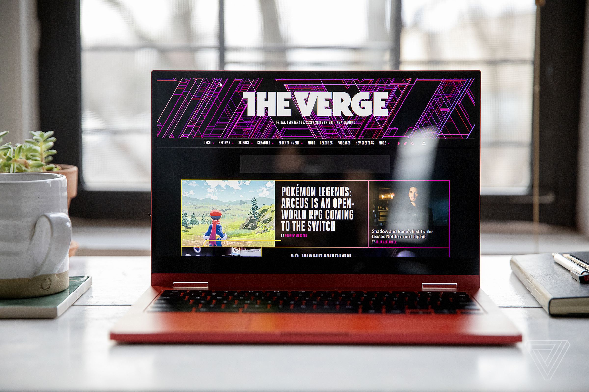 The Samsung Galaxy Chromebook 2 on a desk in front of a window, open with a mug and small plant on its left and some books on its right. The screen displays The Verge homepage.
