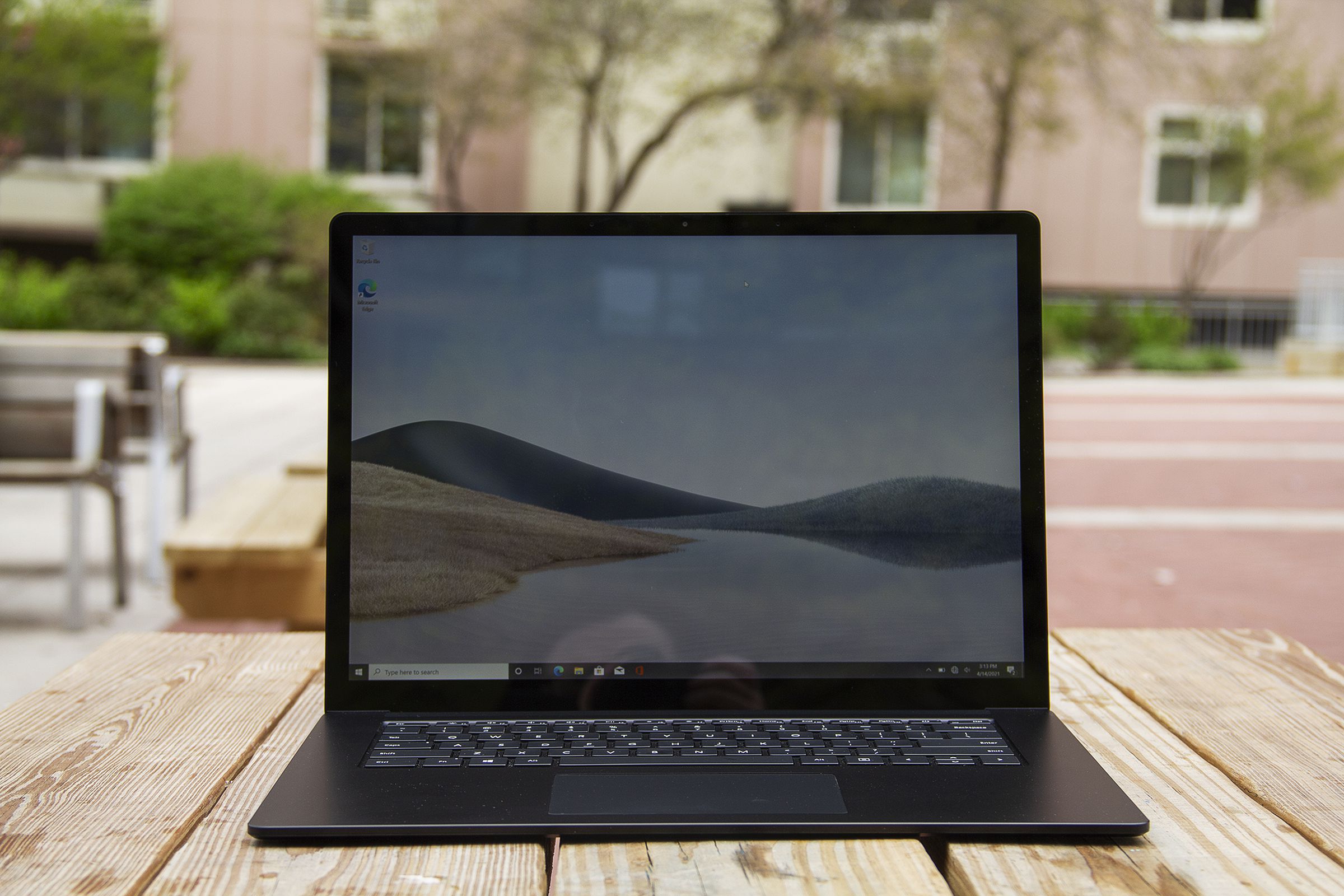 The Surface Laptop 4 15-inch open on a picnic table with a pink apartment building in the background. The screen displays sand dunes with a blue sky.