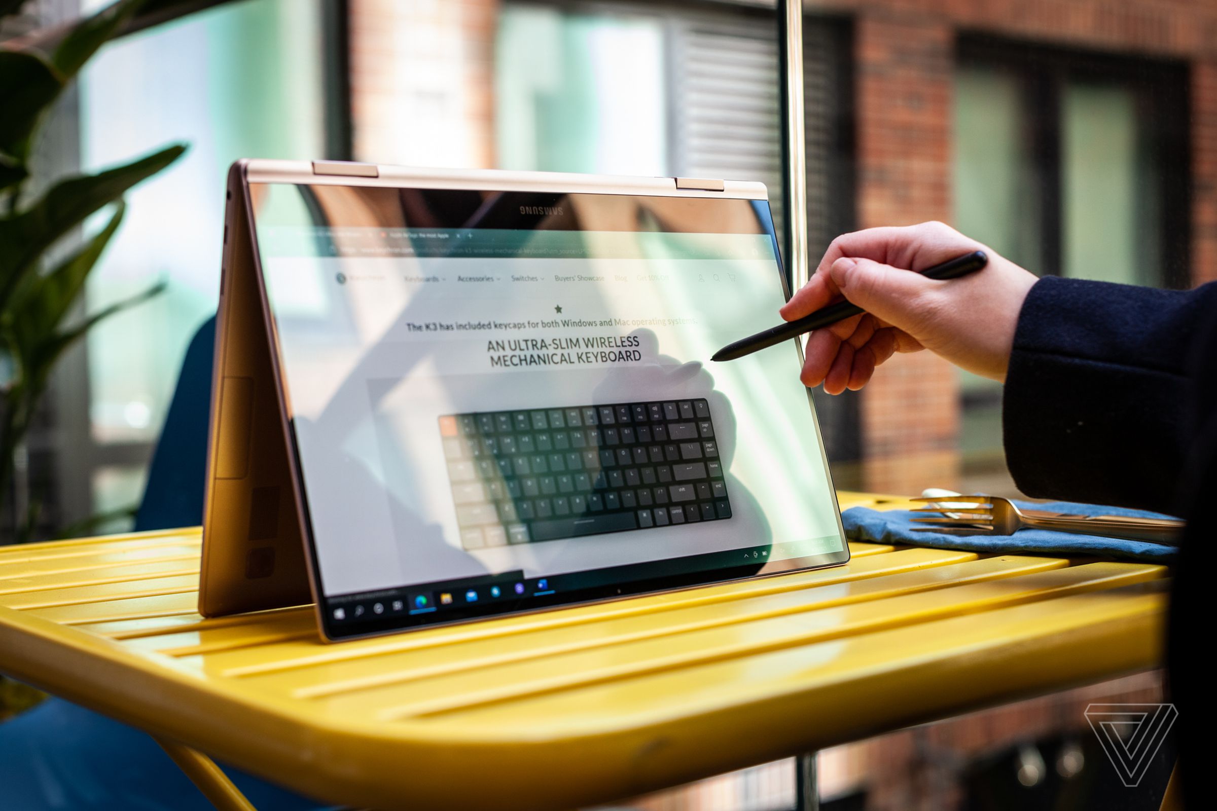 A user uses the Galaxy Book Pro 360 in tent mode on an outdoor picnic table with the S-Pen. The screen displays a mechanical keyboard review on The Verge.