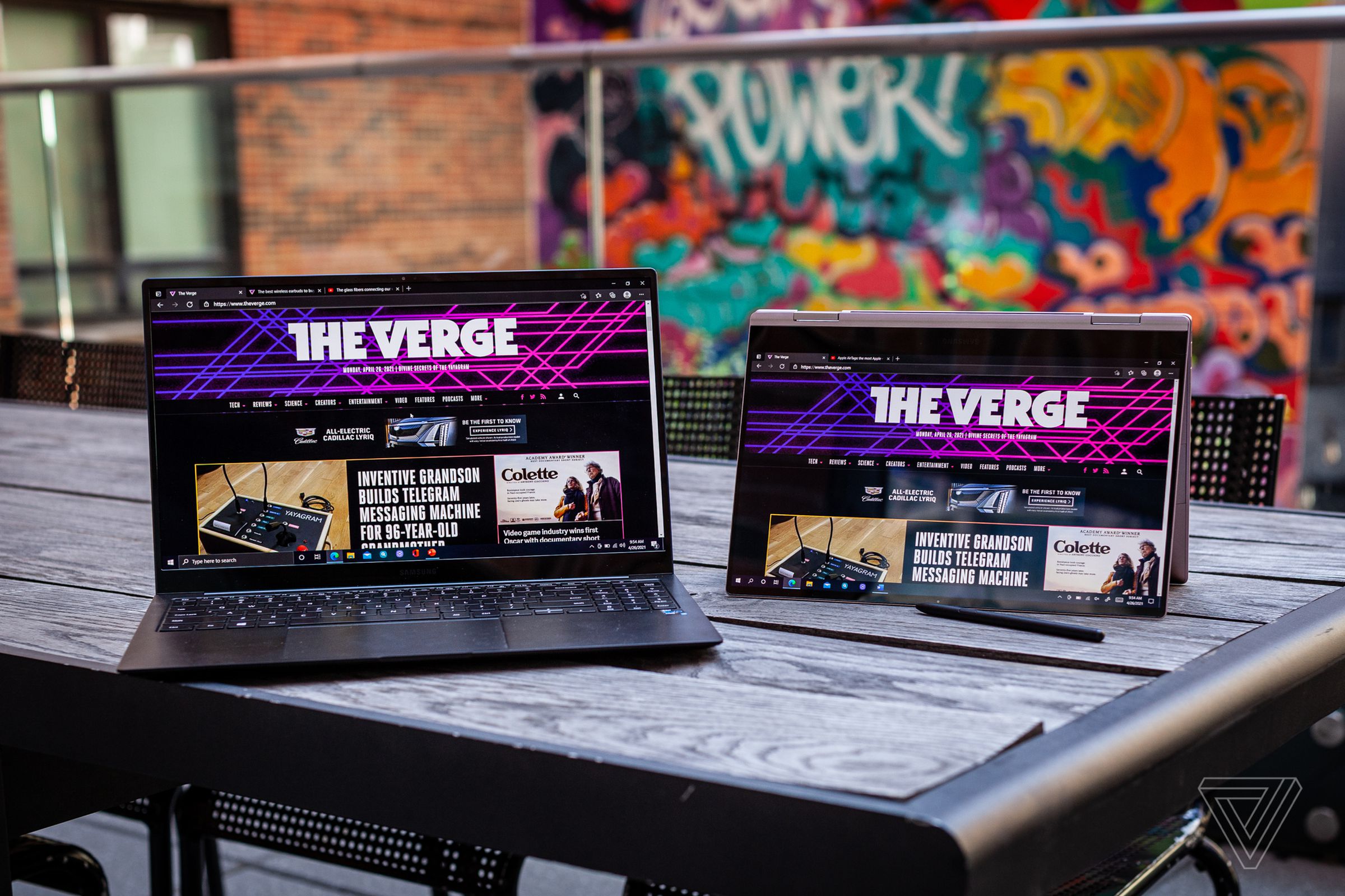 The Samsung Galaxy Book Pro open, and the Samsung Galaxy Book Pro 360 in tent mode, side by side on a picnic table. Both screens display The Verge homepage. The S-Pen lies in front of the Galaxy Book pro 360.