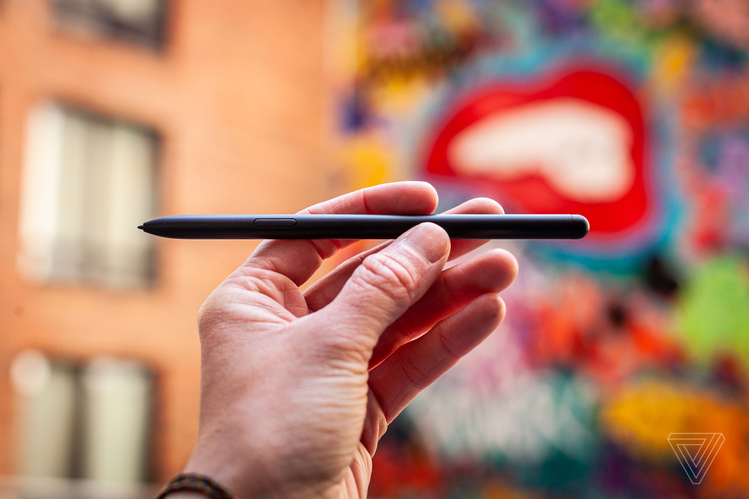 A hand holds the S-Pen in front of a colorful wall.
