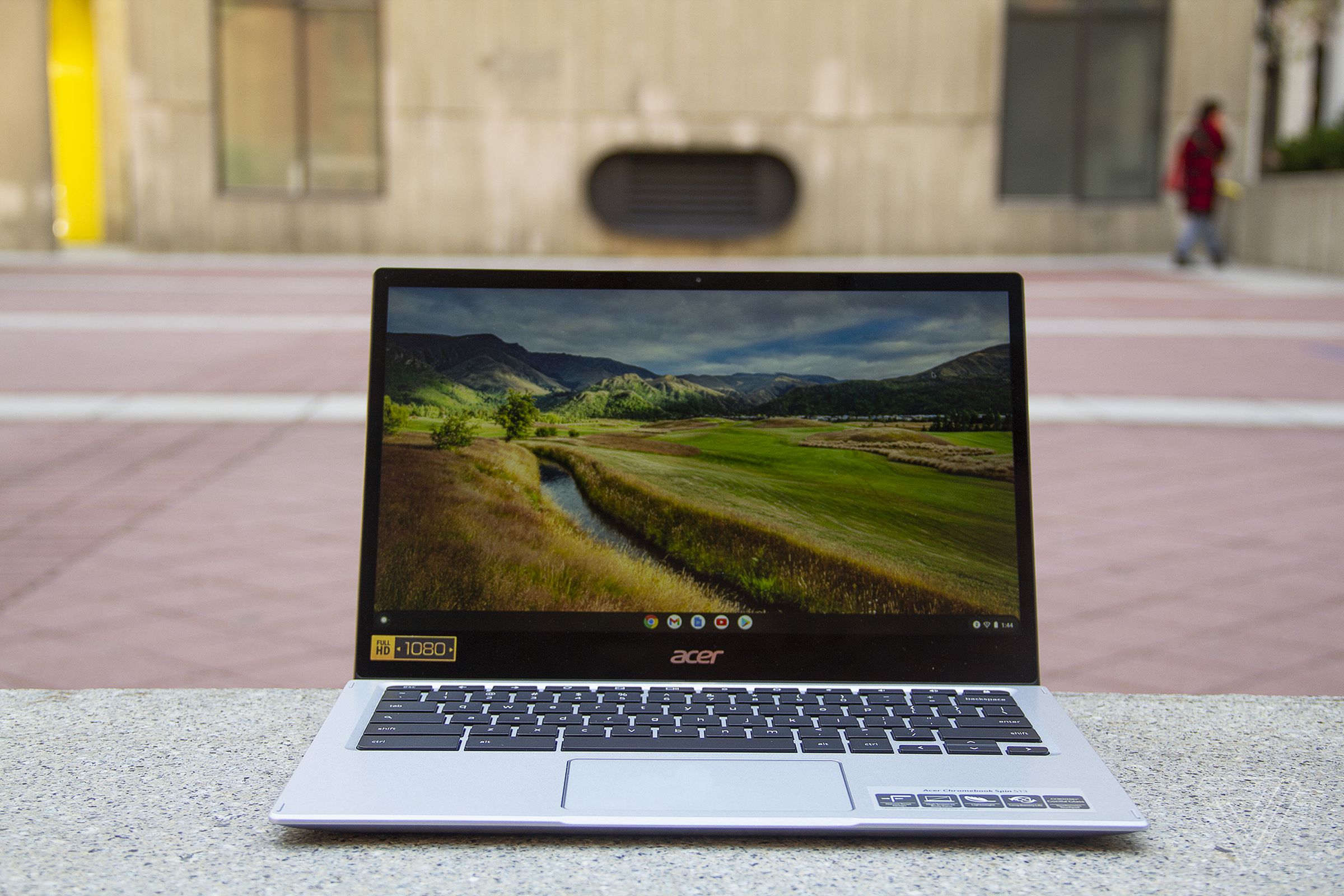 The Acer Chromebook Spin 513 sits on a stone bench, open. The screen displays a pastoral scene.