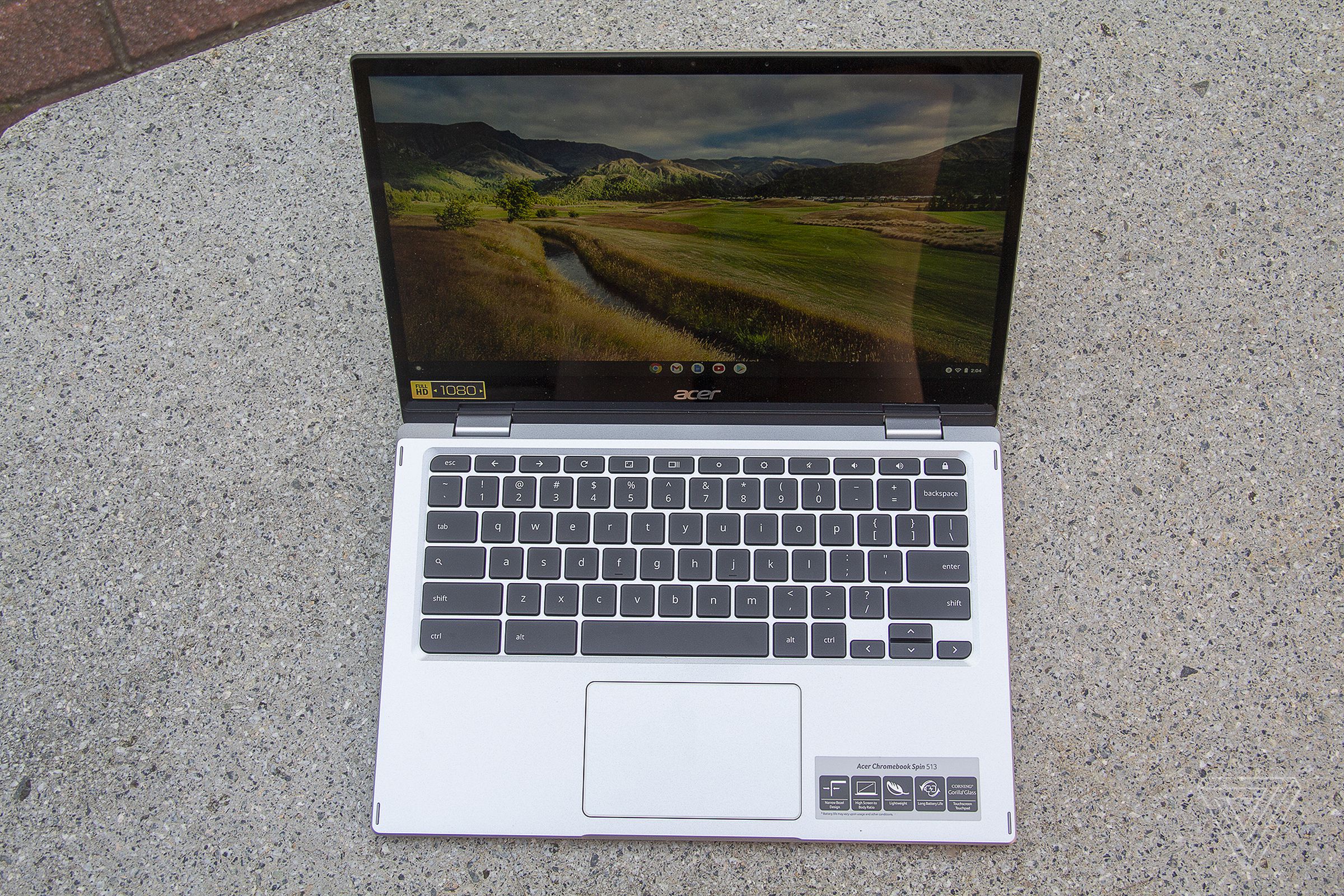 The Acer Chromebook Spin 513 on a stone bench, seen from above, open. The screen displays a pastoral landscape.