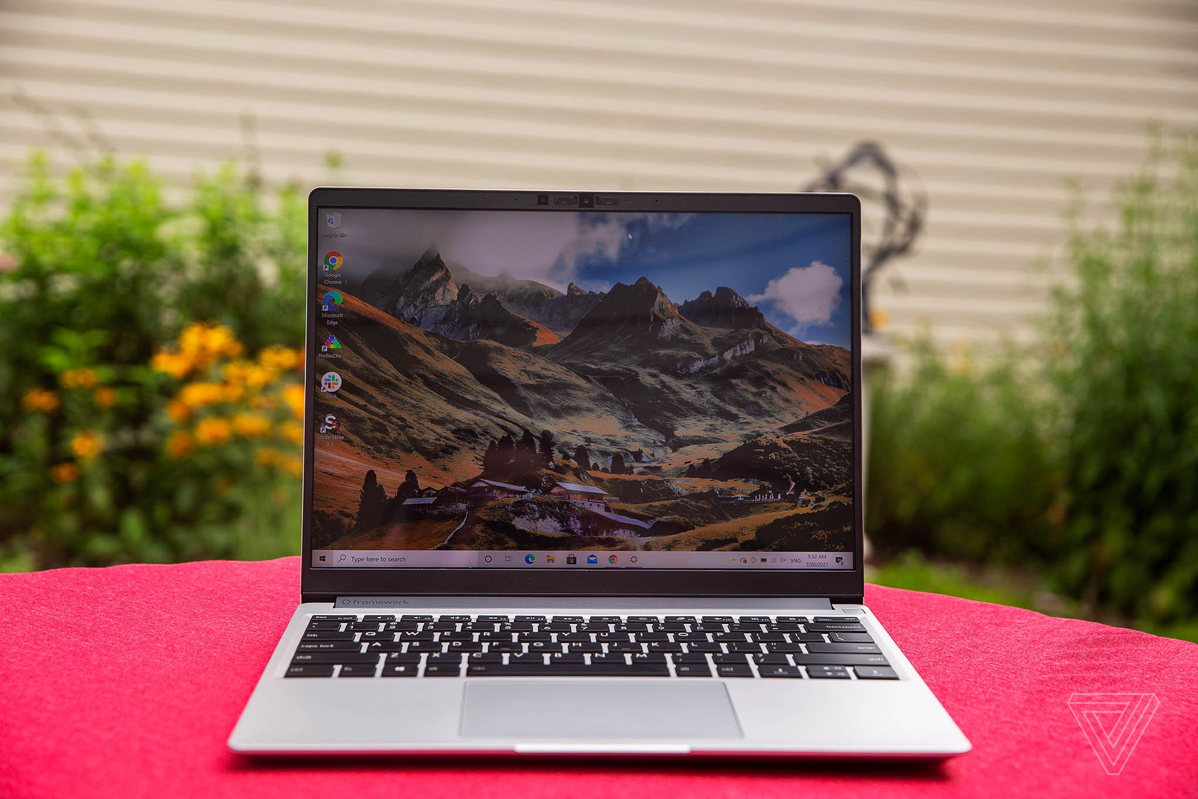 The Framework laptop open, outdoors, on a red tablecloth with a garden and the wall of a house in the background. The screen displays a mountainous landscape.