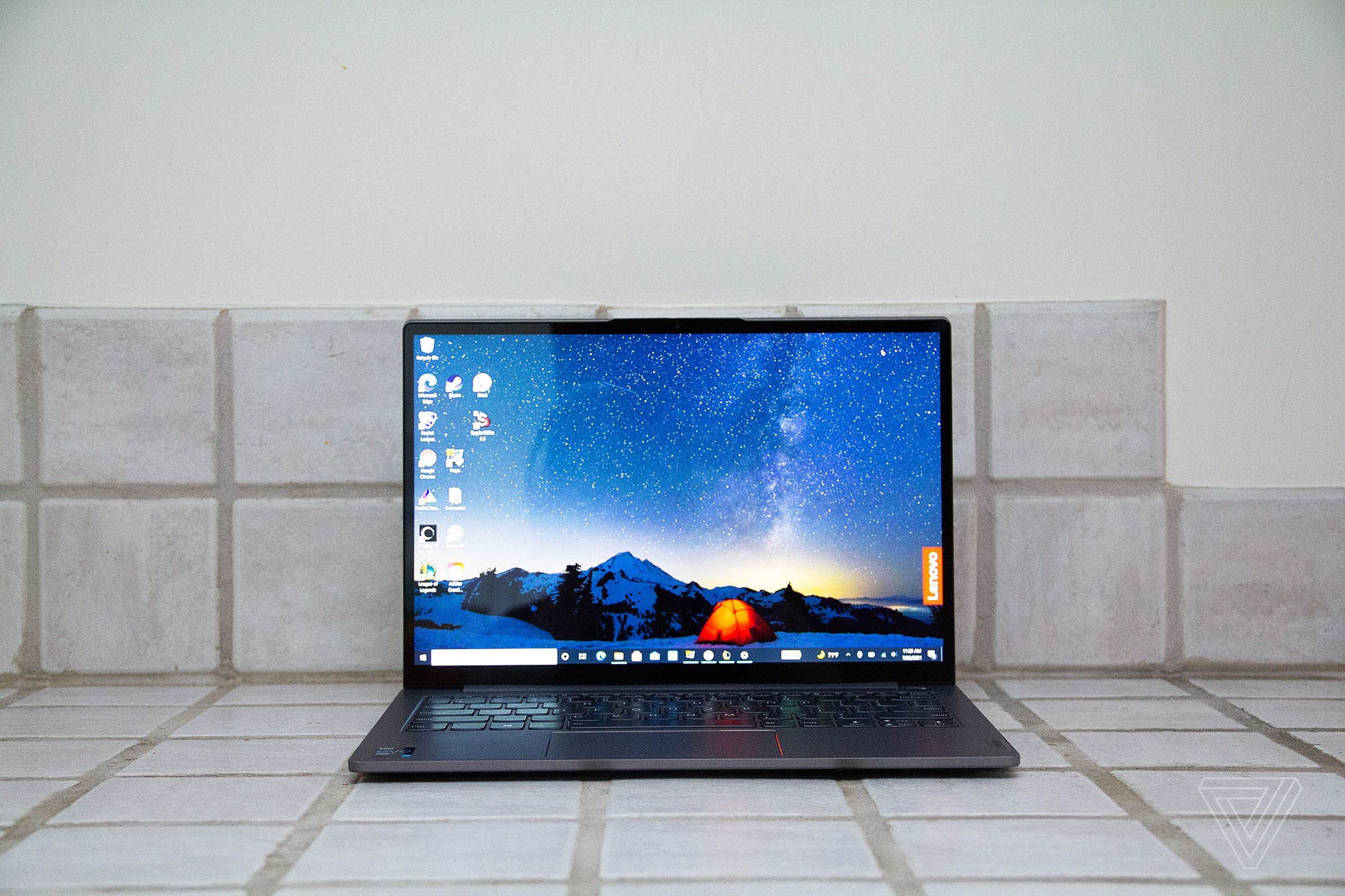 The Lenovo ThinkBook 13s seen from the front on a white-tiled countertop. The screen displays a night mountain scene with a bright tent in the center and a Lenovo banner on the right side.