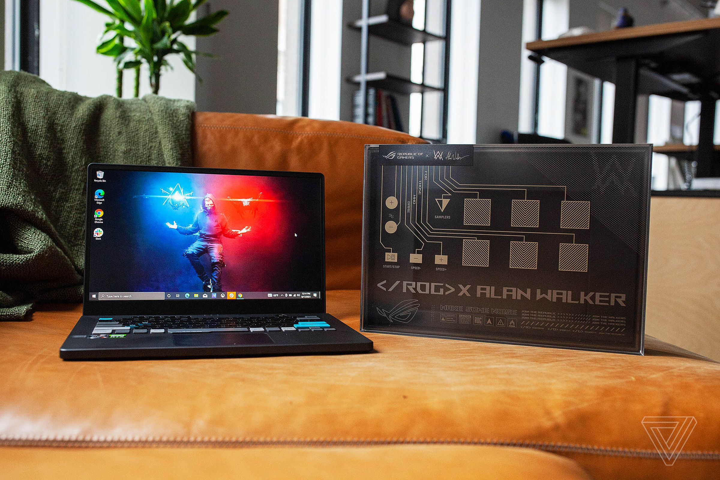 The Asus ROG Zephyrus G14 Alan Walker Edition on a couch seen from the front, open. The screen displays Alan Walker hovering on a red and blue background, conjuring lights of each color with his hands. To its left is its box, with 11 DJ buttons and ROG X ALAN WALKER printed on the bottom.