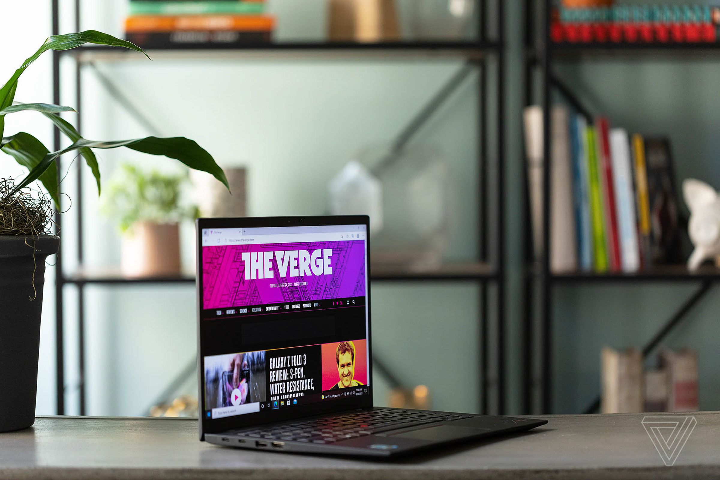 The Lenovo ThinkPad X1 Carbon gen 9 on a table open, angled to the right. The screen displays The Verge homepage. In the background is a bookshelf with potted plants and books.
