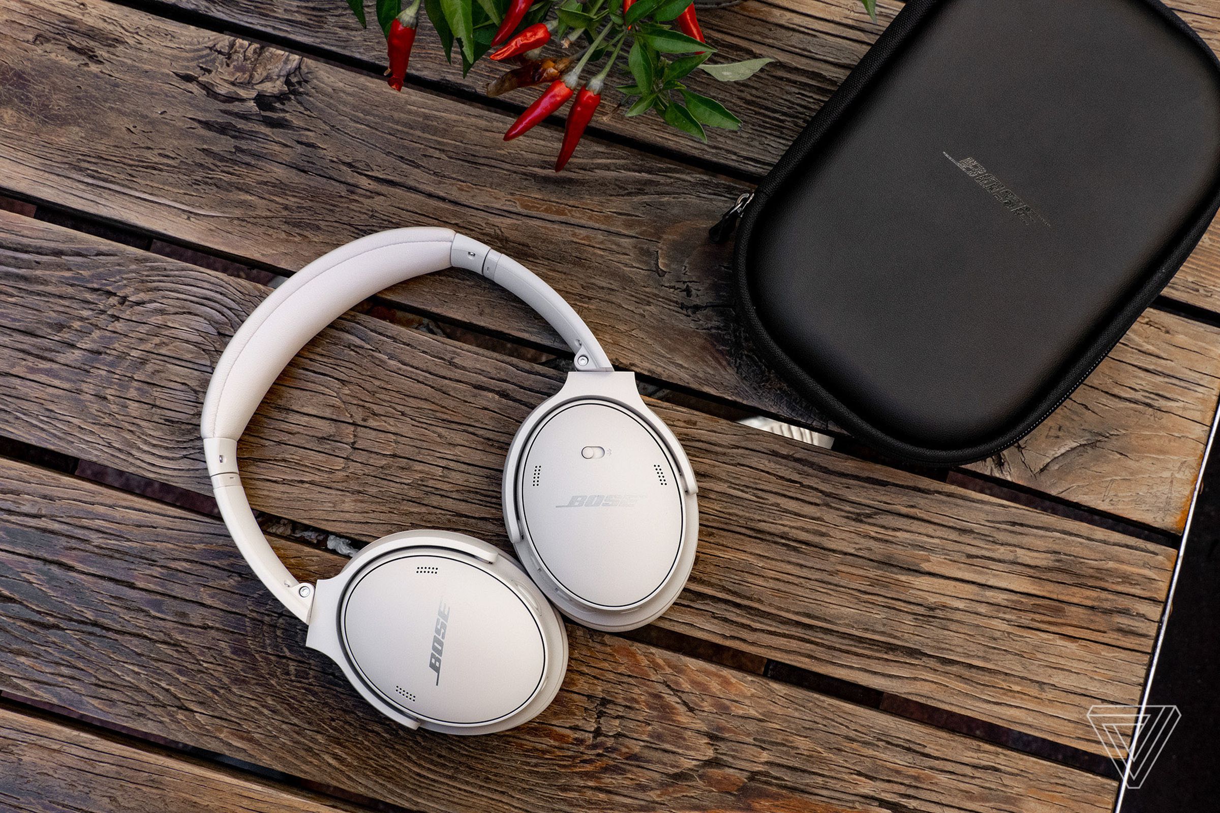 The white version of the Bose QuietComfort 45 headphones on a wood picnic table with their black protective zipper case beside them.