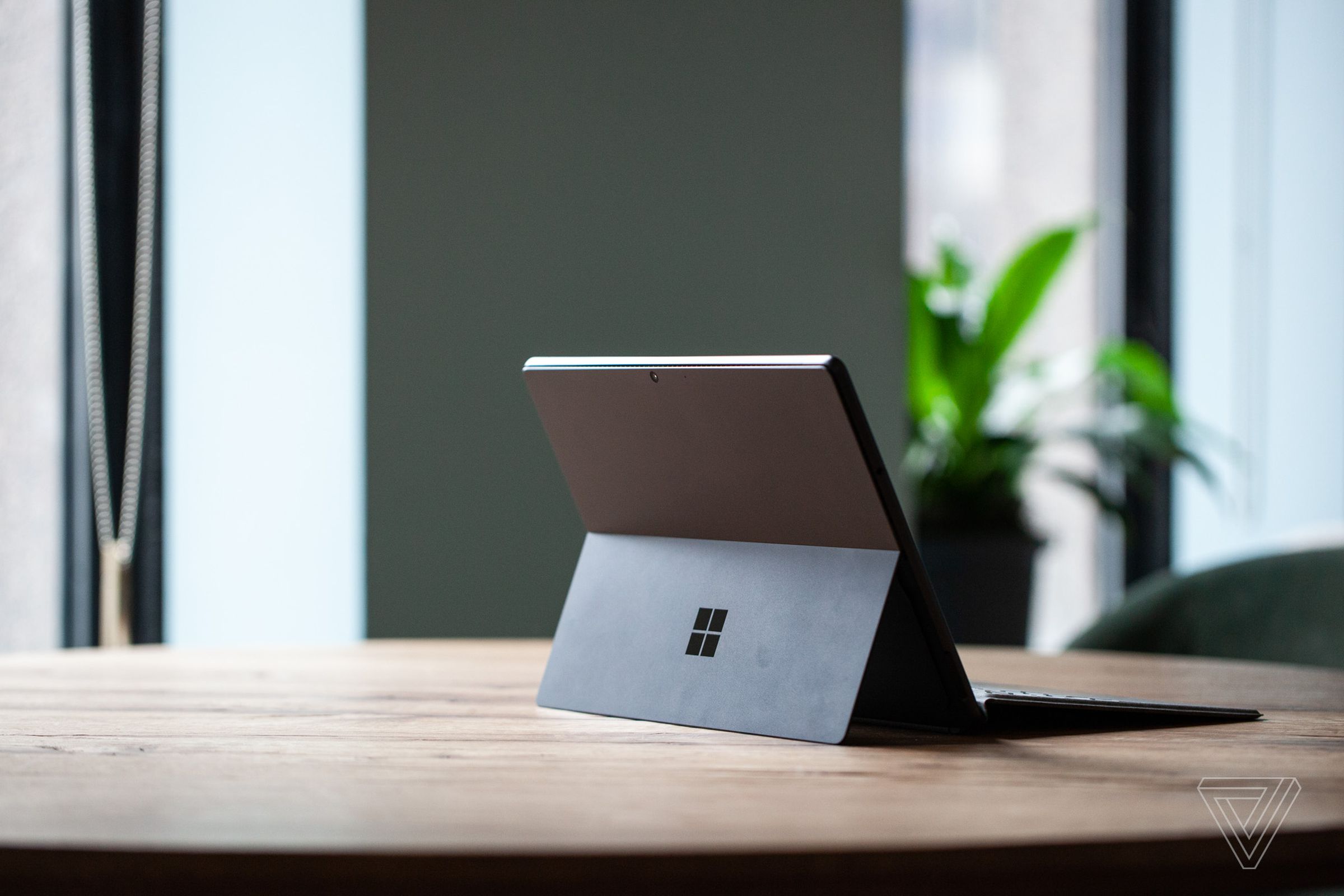The Surface Pro 8 on a table seen from the back to the right, open, with the Signature Keyboard attached and the kickstand out. In the background is a green wall and a potted plant.