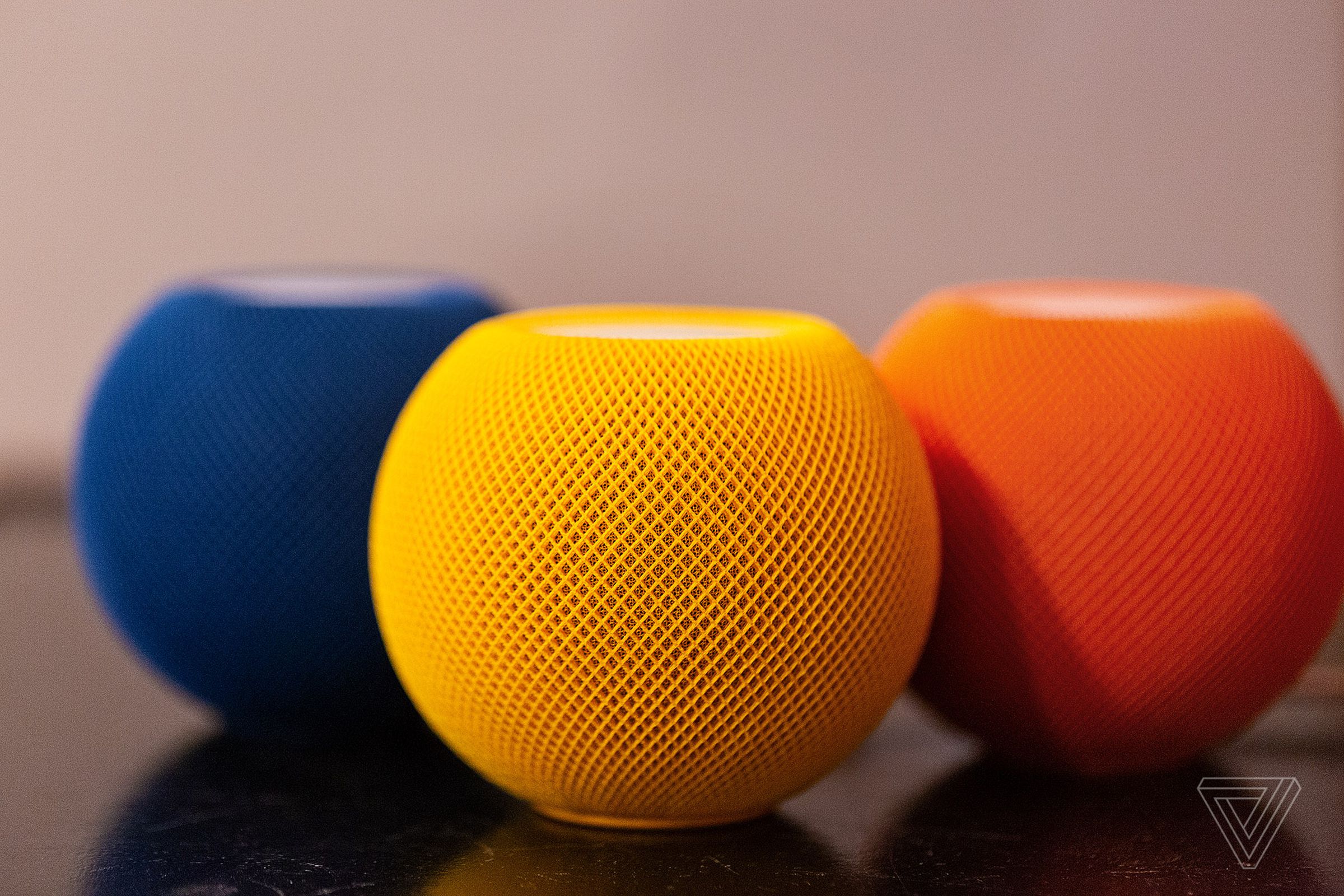 Three of Apple’s HomePod Mini smart speakers standing on a desk in various shades including orange, yellow, and blue.