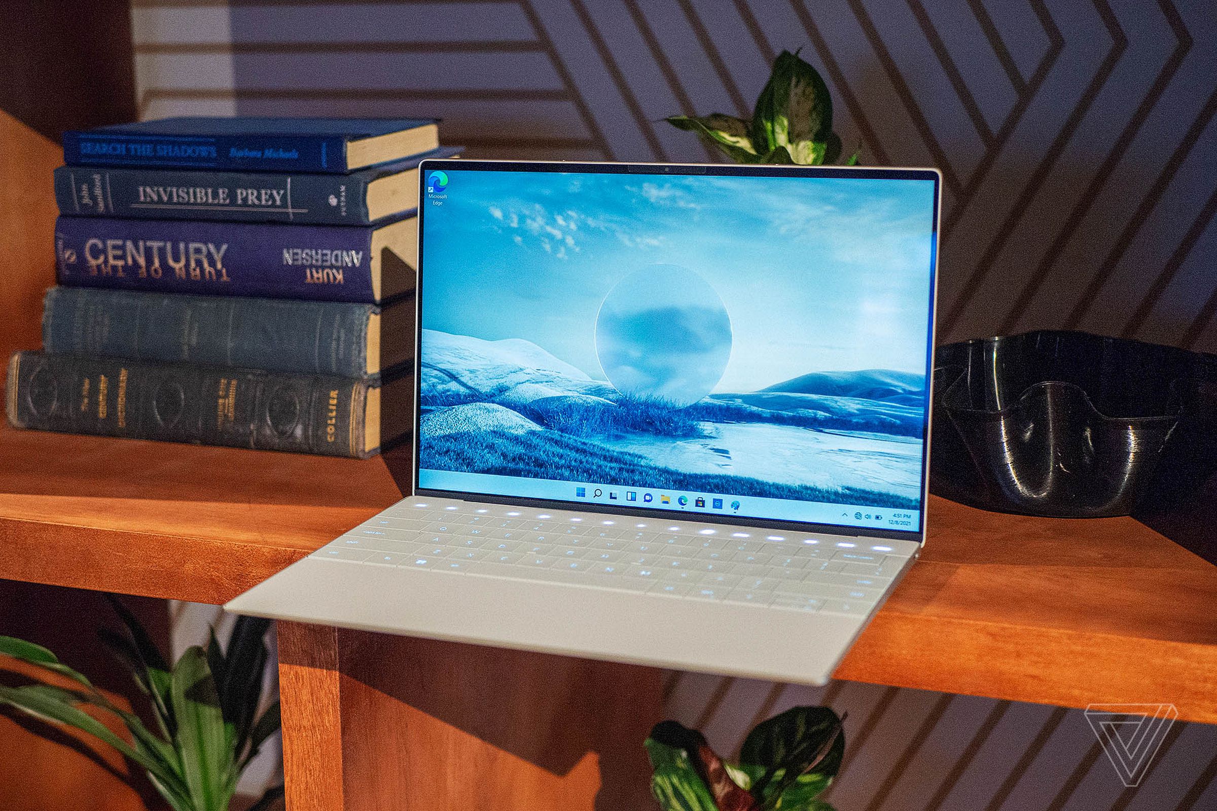 The Dell XPS 13 Plus open and angled to the left on a shelf. The screen displays a blue arctic setting. A pile of books is to its right.