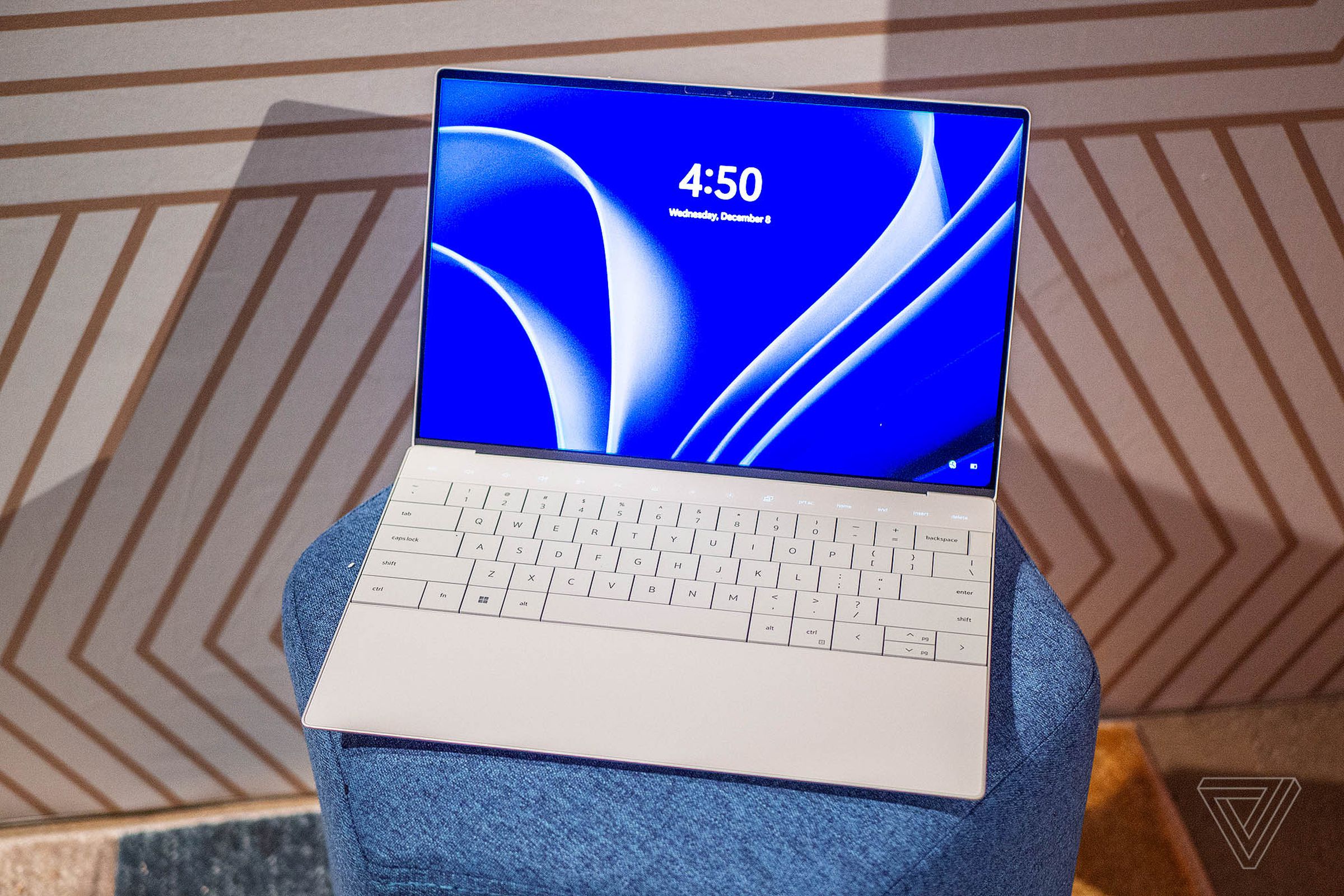 The Dell XPS 13 Plus seen from above on a blue plush stool. The screen displays a blue and white background and the time 4:50.