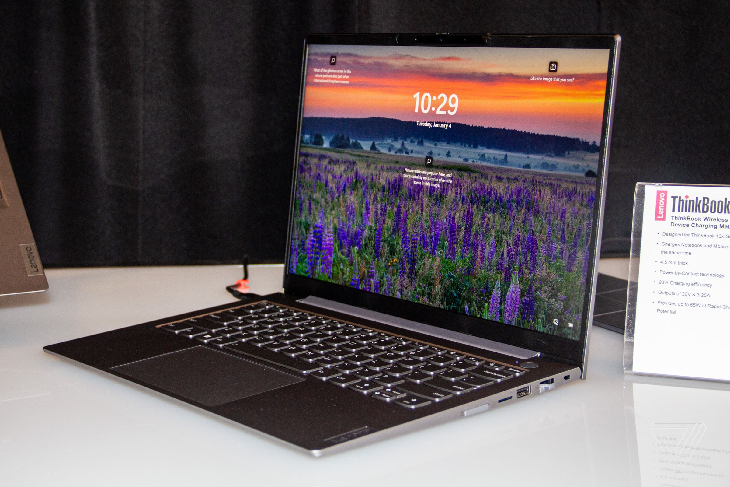 The Lenovo ThinkBook 14 Gen 4 open on a white table, plugged in, angled to the left. The screen displays a field of purple flower beneath an orange sky.