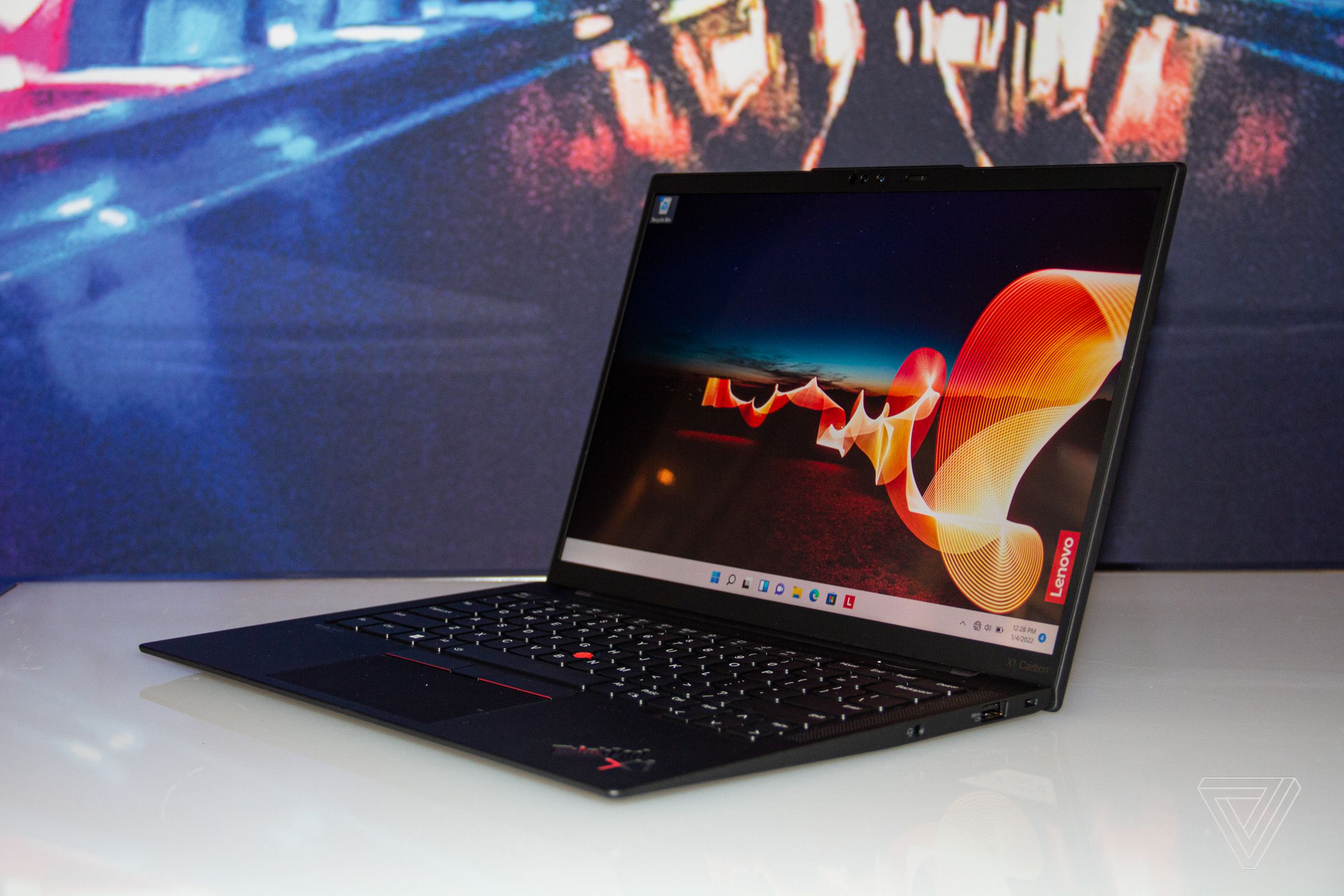 The ThinkPad X1 Carbon open on a white table angled to the left. The screen displays a night desert setting with an orange ribbon running through it and the Lenovo logo on the right side.