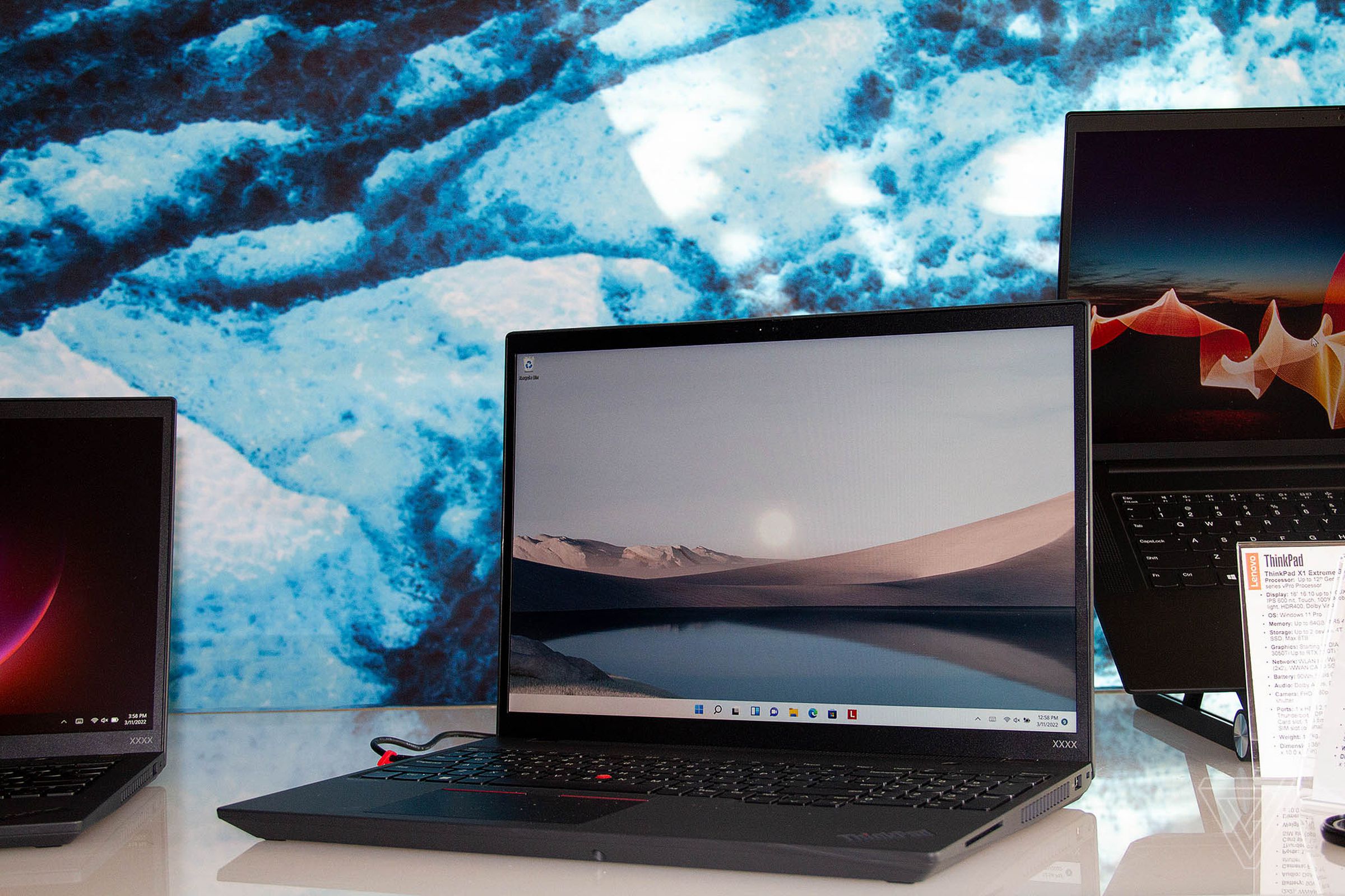 The Lenovo ThinkPad T16 on a white table open, angled to the left. The screen displays a mountain range from across a lake.
