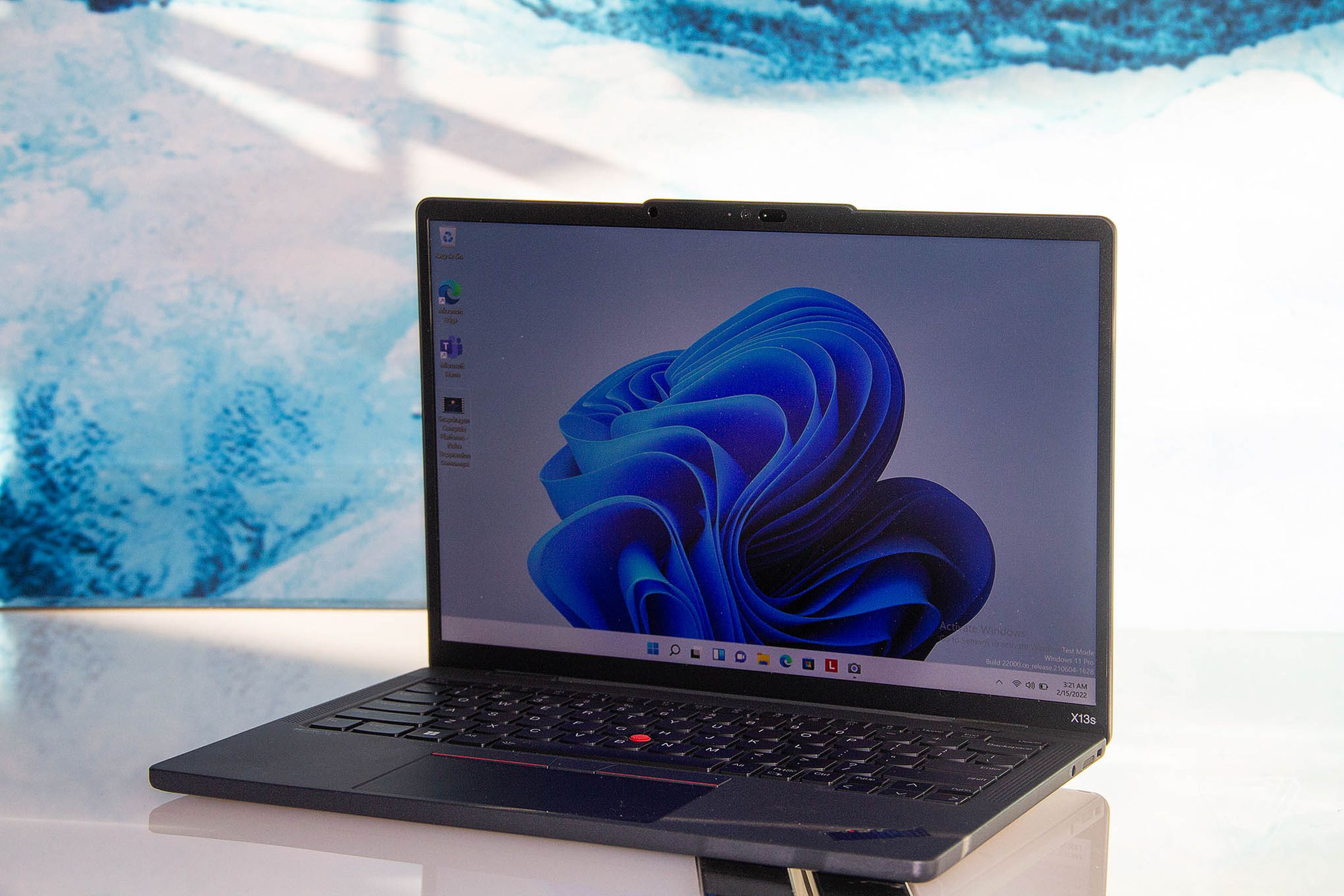 The Lenovo ThinkPad X13s open on a white table angled to the left. The screen displays a blue ribbon pattern on a light blue background.