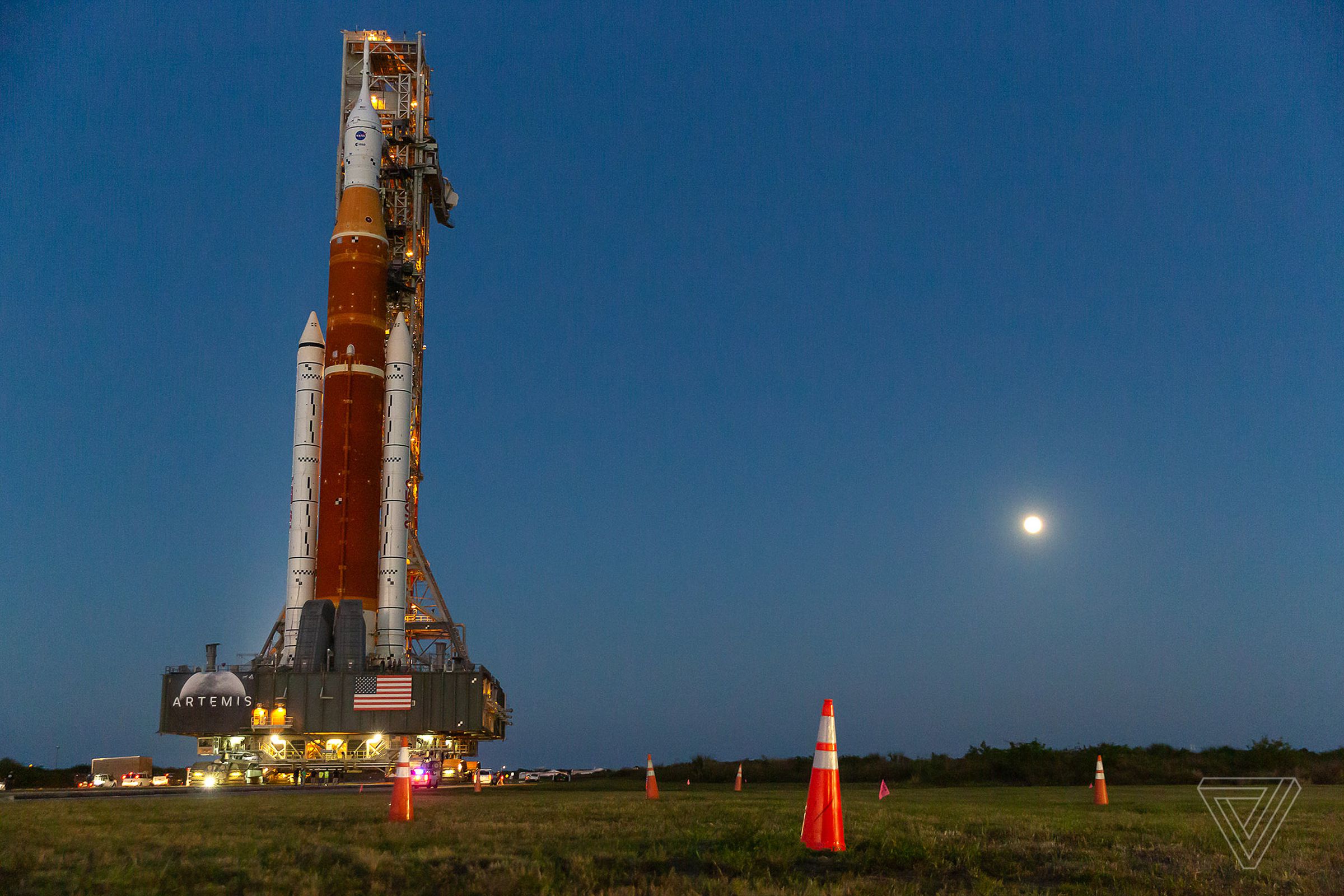 The Moon, the eventual destination of the SLS, shines in the background as the rocket rolls out.