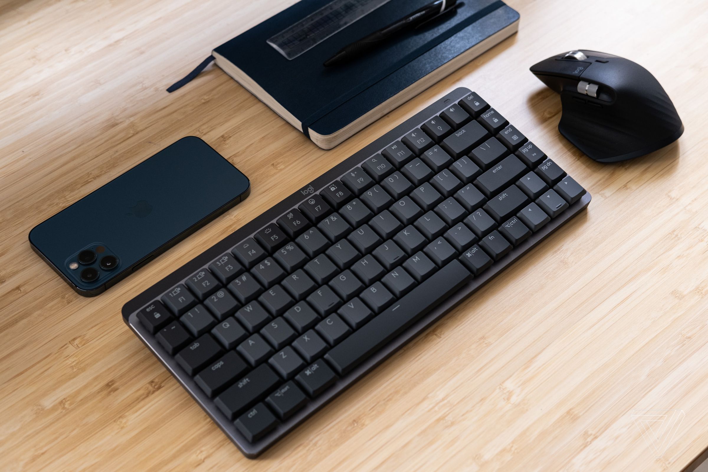 A Logitech MX Mechanical Mini keyboard on a wood desk, beside a mouse, phone, and notebook.