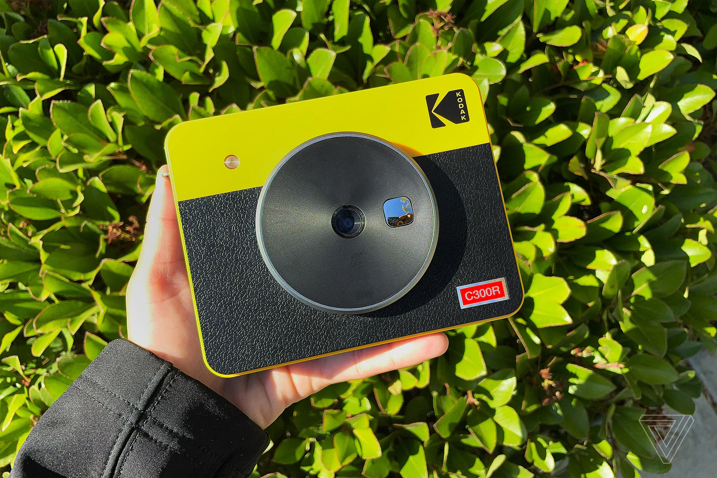 The yellow and black Kodak Mini Shot 3 Retro being held up sideways with a hand and a bush as the background.