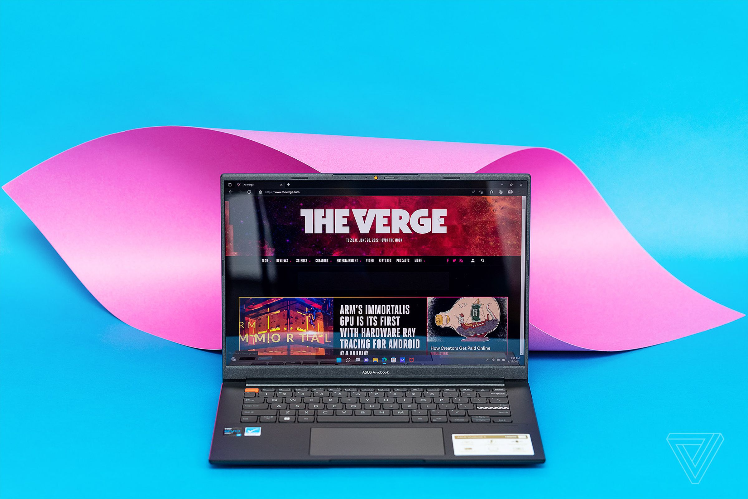 The Asus Vivobook S 14X seen from above on a blue and pink surface. The screen displays The Verge homepage.