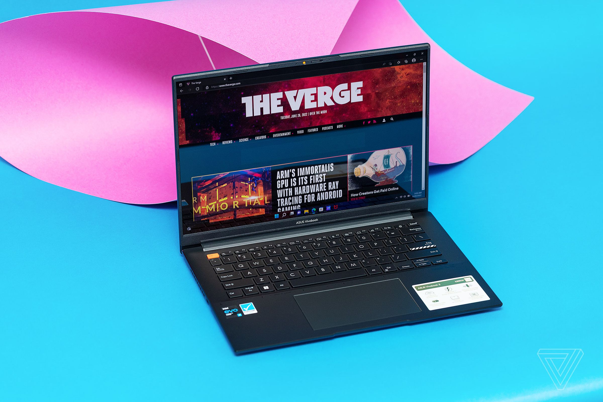 The Asus Vivobook S 14X OLED seen from above on a blue and pink background. The screen displays The Verge homepage.