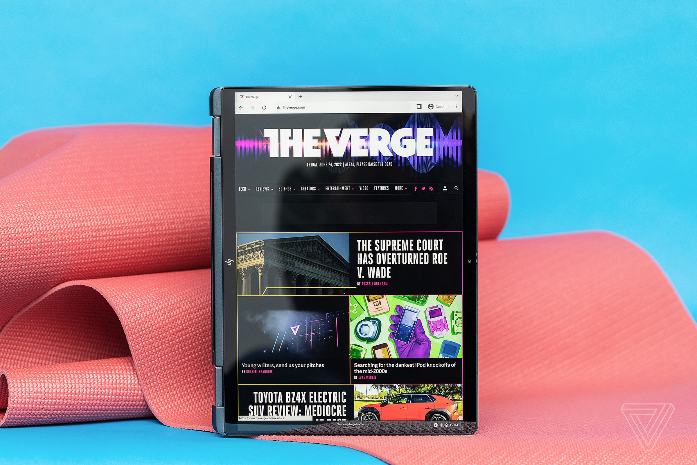 The HP Dragonfly Chromebook in tablet mode on a blue and pink background. The screen displays The Verge homepage.