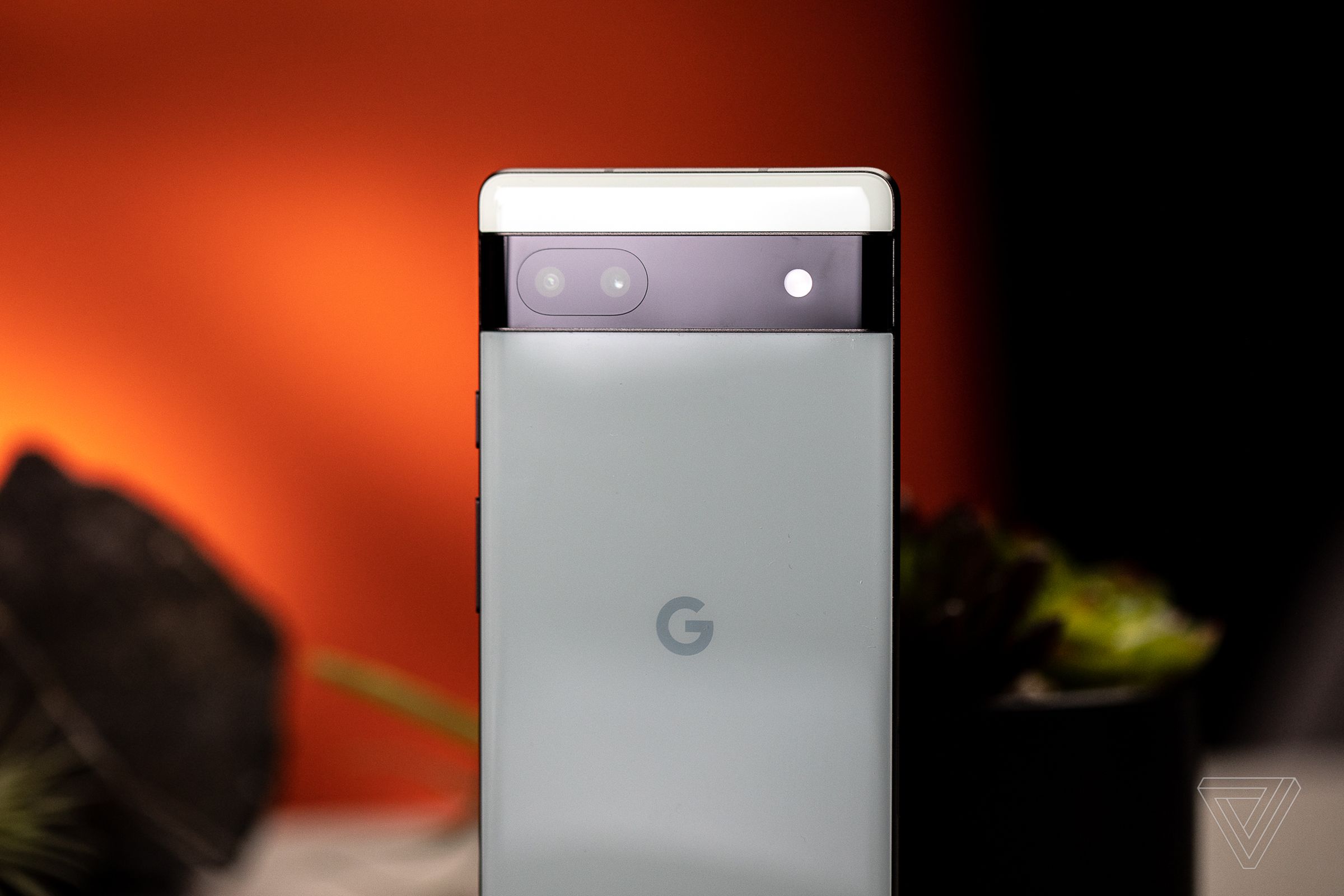 A close-up photo of the top three-quarters of the Pixel 6A’s rear. The phone is light green with a black camera bar across it. The background is a burnt orange with small plant life behind the phone.