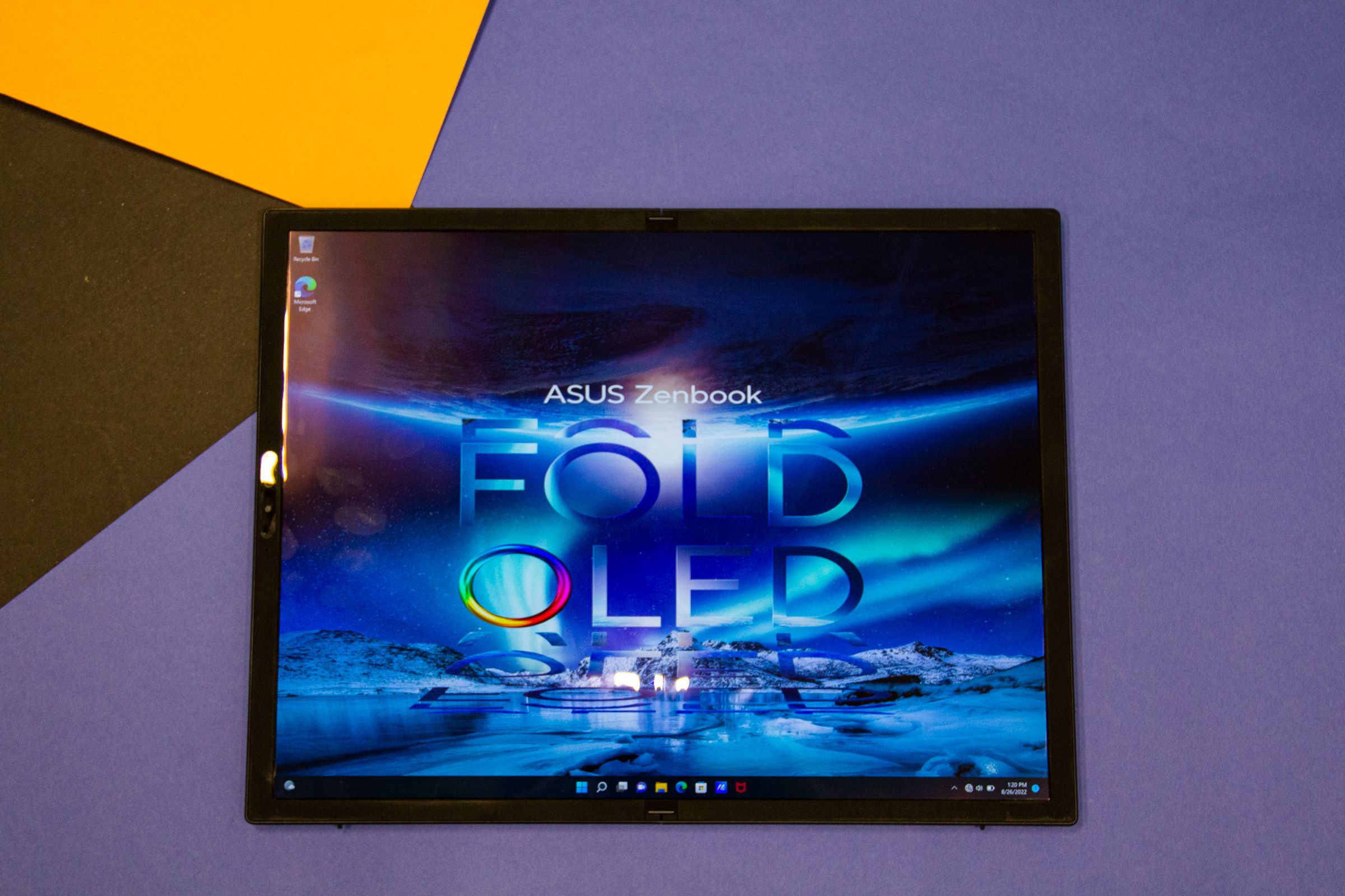 The Asus Zenbook Fold OLED in laptop mode laid out on a table. The screen displays the Asus Zenbook Fold OLED logo on a blue background.