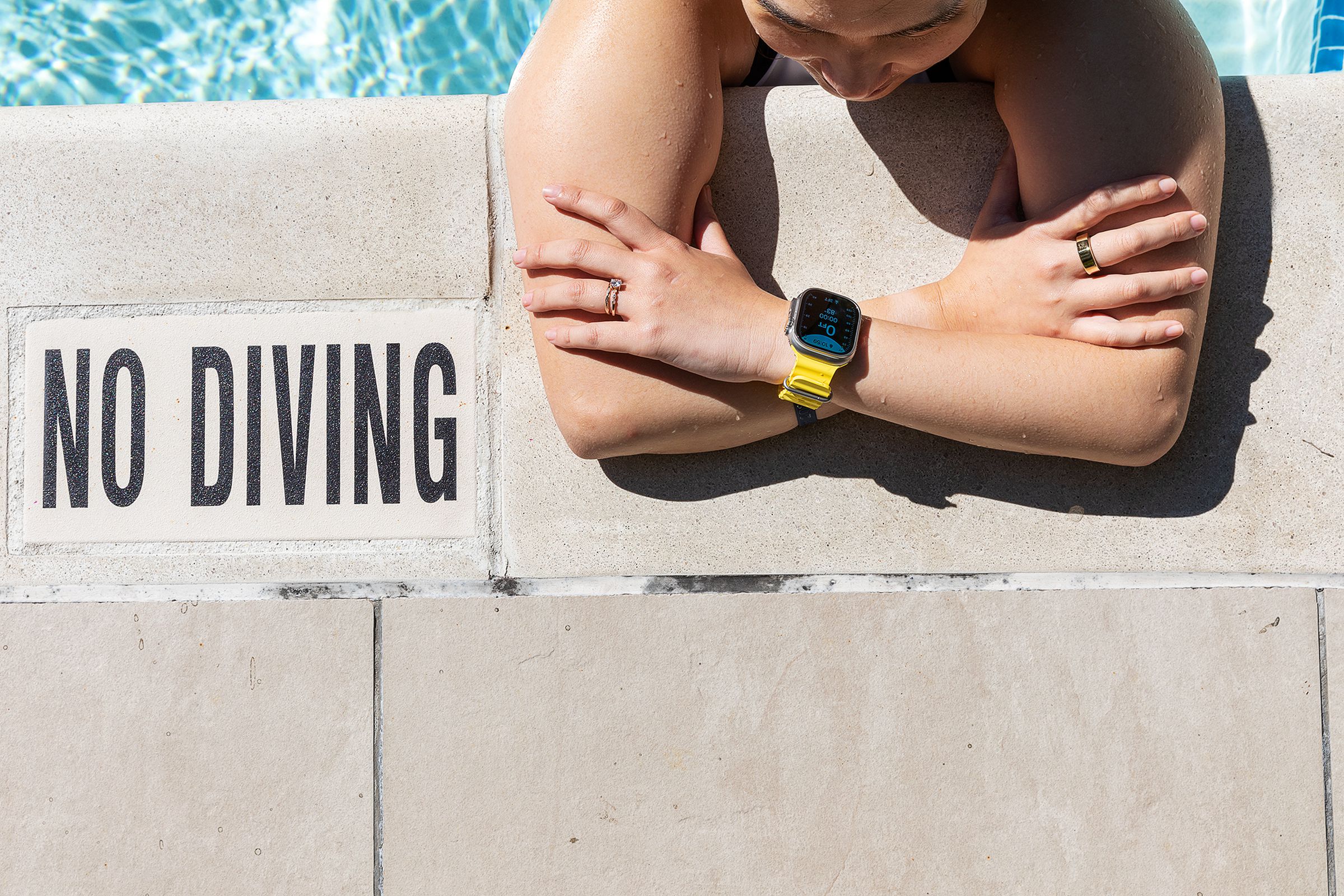 Mulher encostada na beira da piscina usando o Apple Watch Ultra ao lado de uma placa de proibido mergulhar.