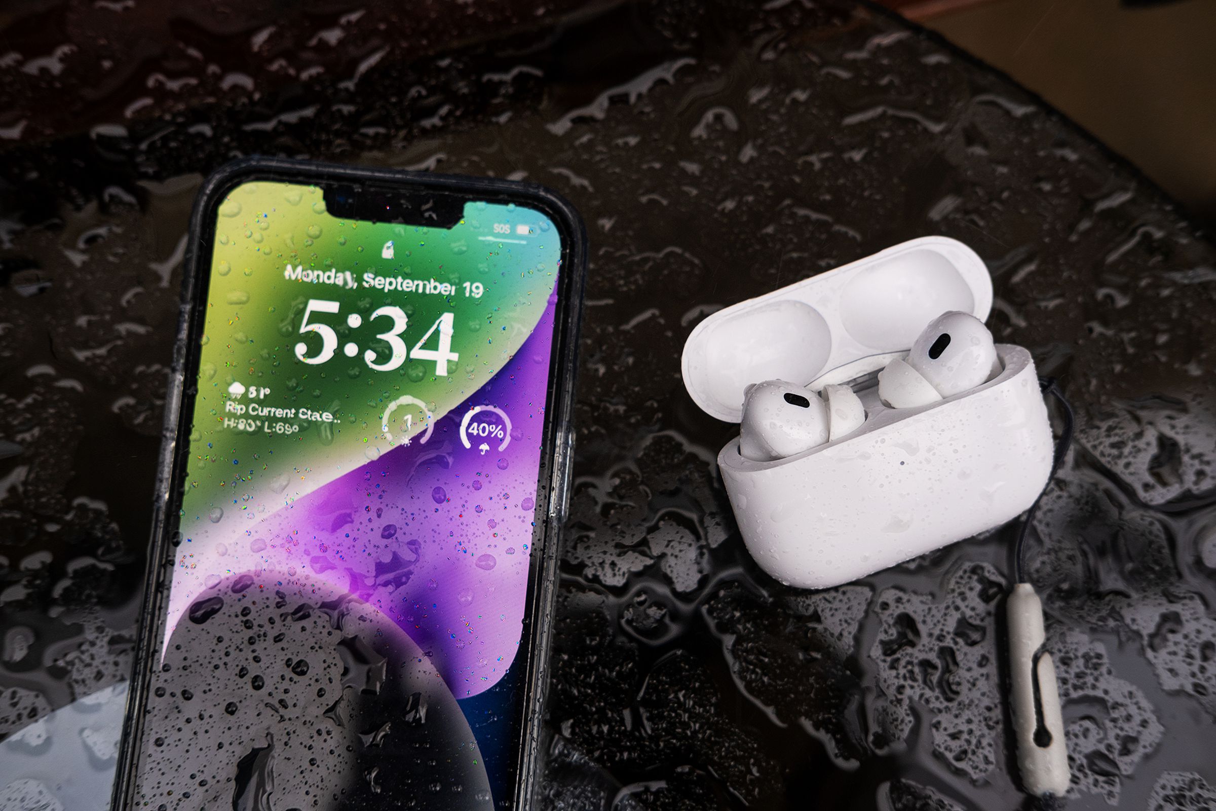 A photo of the iPhone 14 next to Apple’s second-generation AirPods Pro. Both devices are wet with visible rain drops on them.