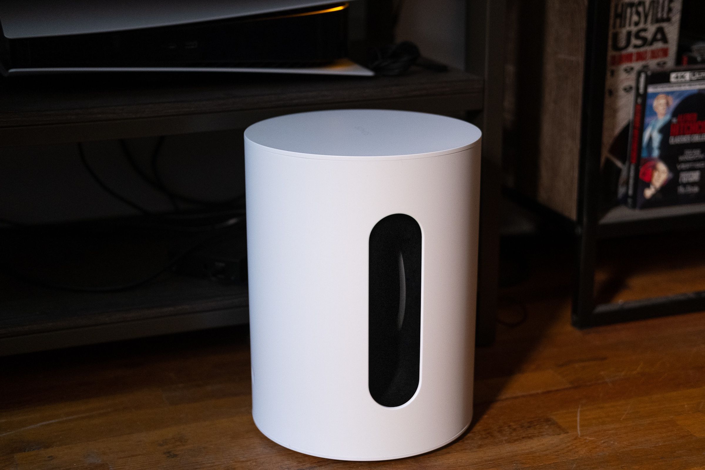 An image of the Sonos Sub Mini pictured on the floor with a TV stand and various components behind it.