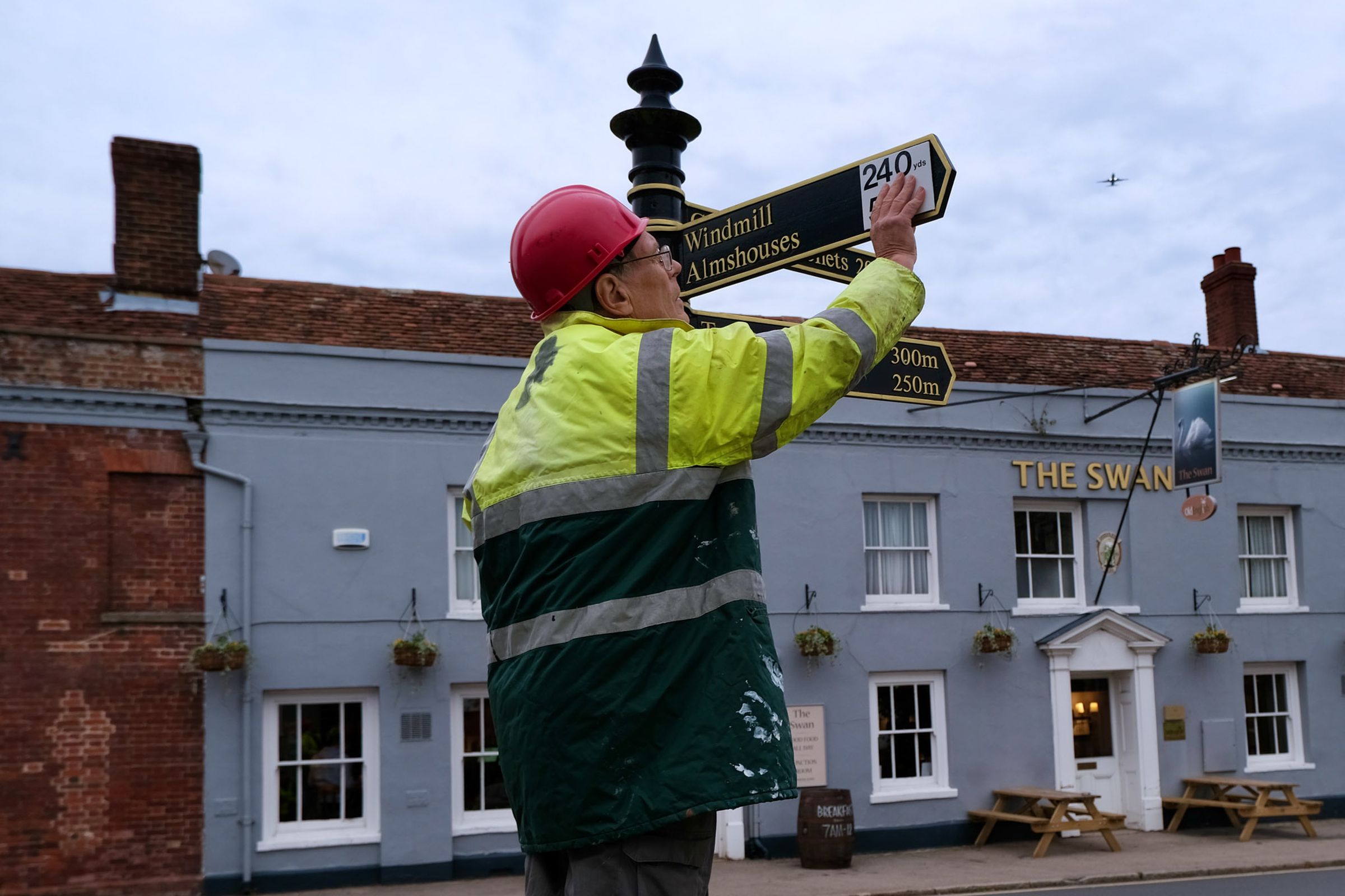 A photo of Tony Bennett placing stickers with new units over the arms of a signpost. He’s standing on a small rickety stepladder and wearing a stained high-visibility jacket and plastic helmet. 