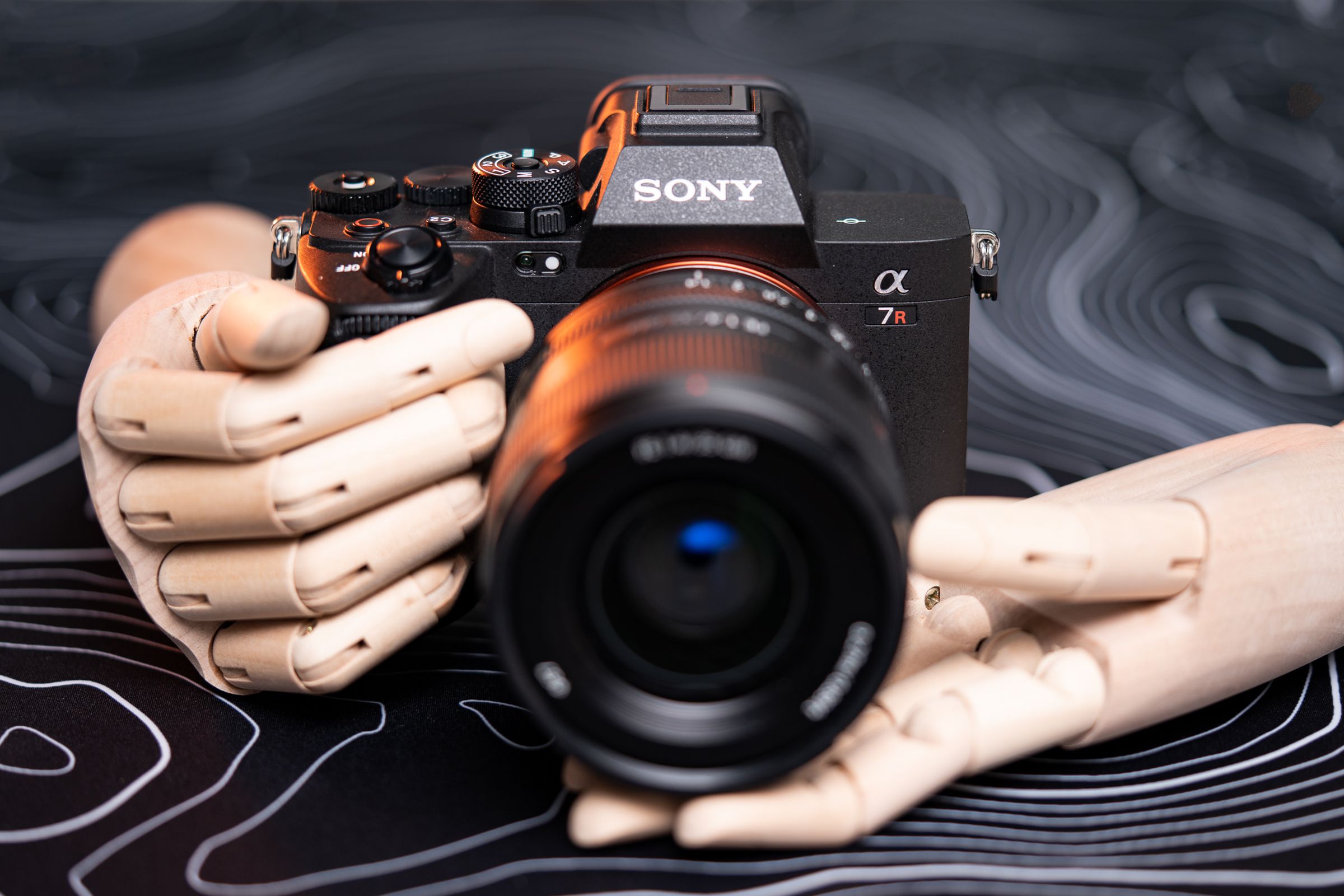 A front view of the Sony A7R V camera with lens attached, sitting on a patterned table with wooden model hands holding it.