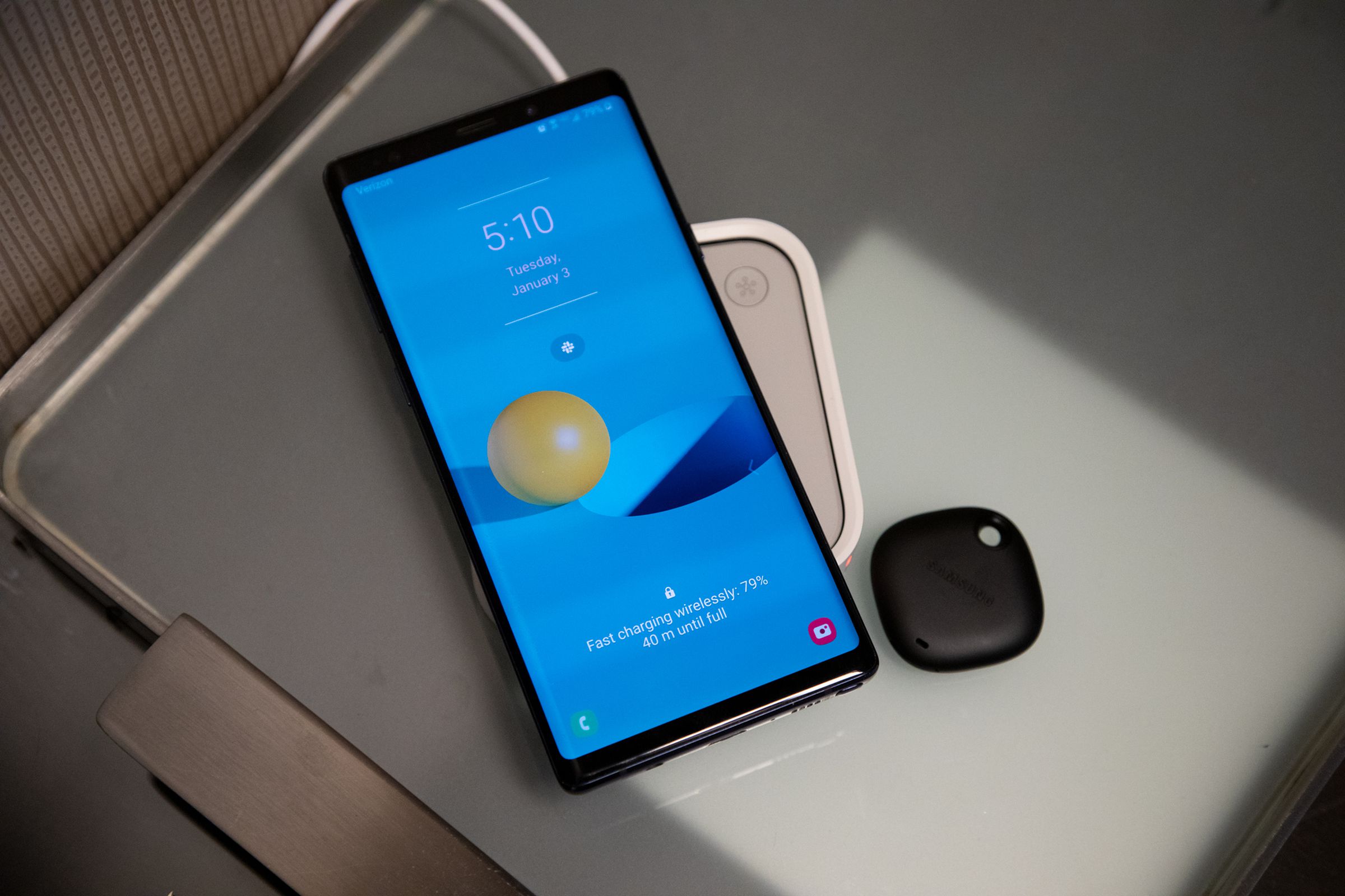 A smartphone on a wireless charger on a nightstand next to a small black tracking device.