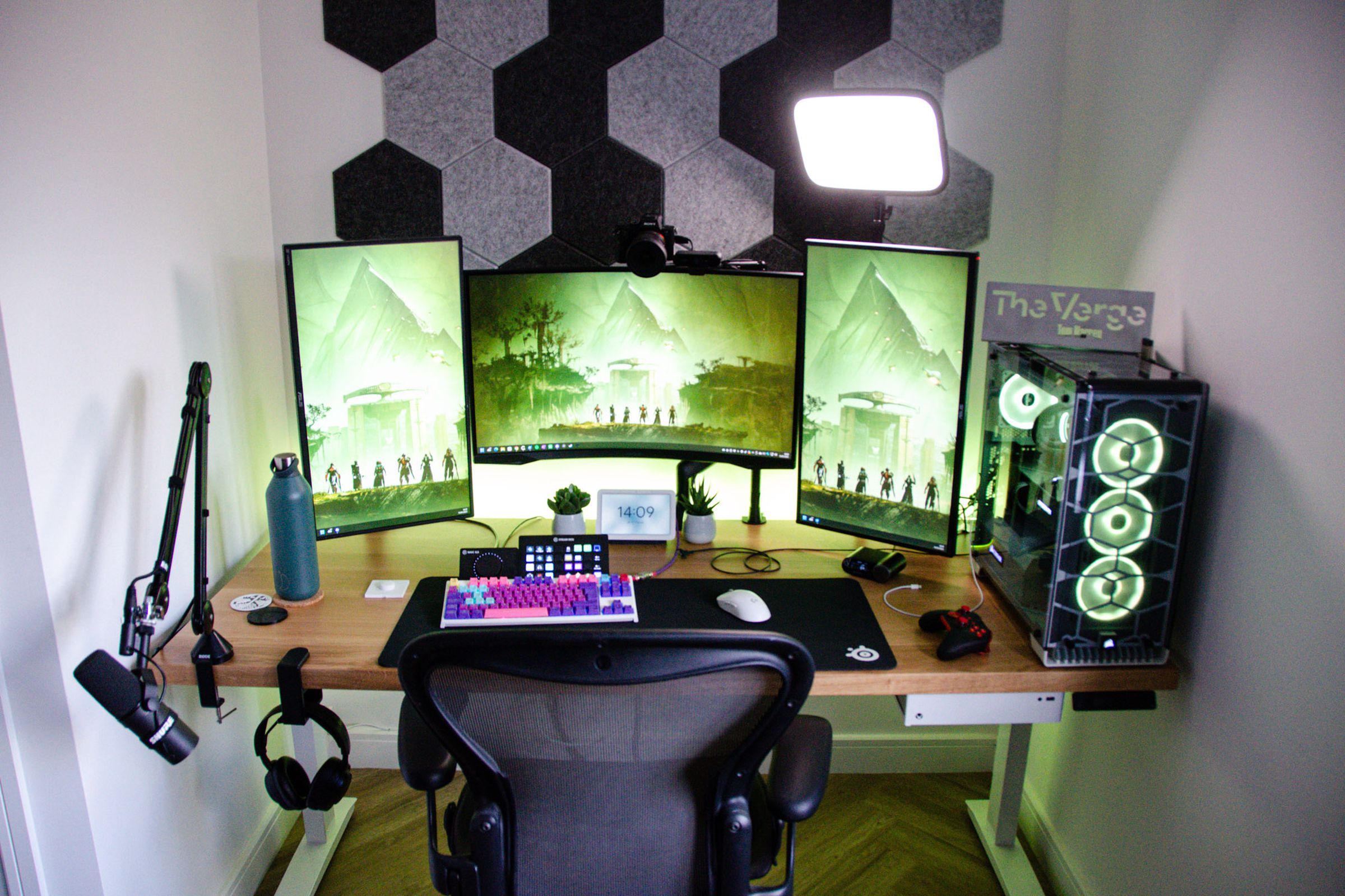 A desk in an alcove with three monitors, a geometrical design on the wall behind it, a colorful gaming keyboard, and a variety of other tech.