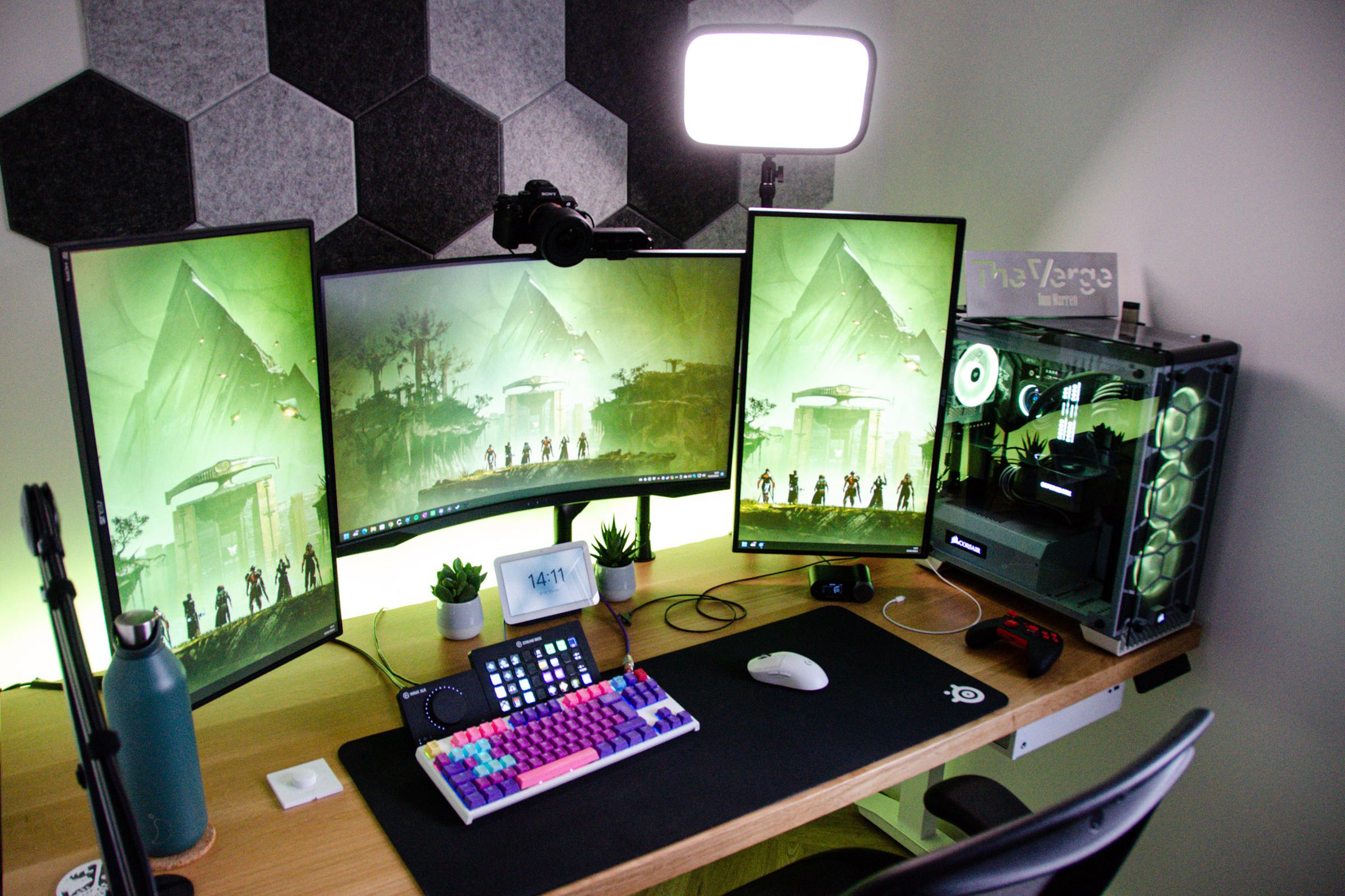 Widok z góry biurka z trzema monitorami, kolorową klawiaturą pośrodku, komputerem po prawej stronie i różnymi innymi urządzeniami.