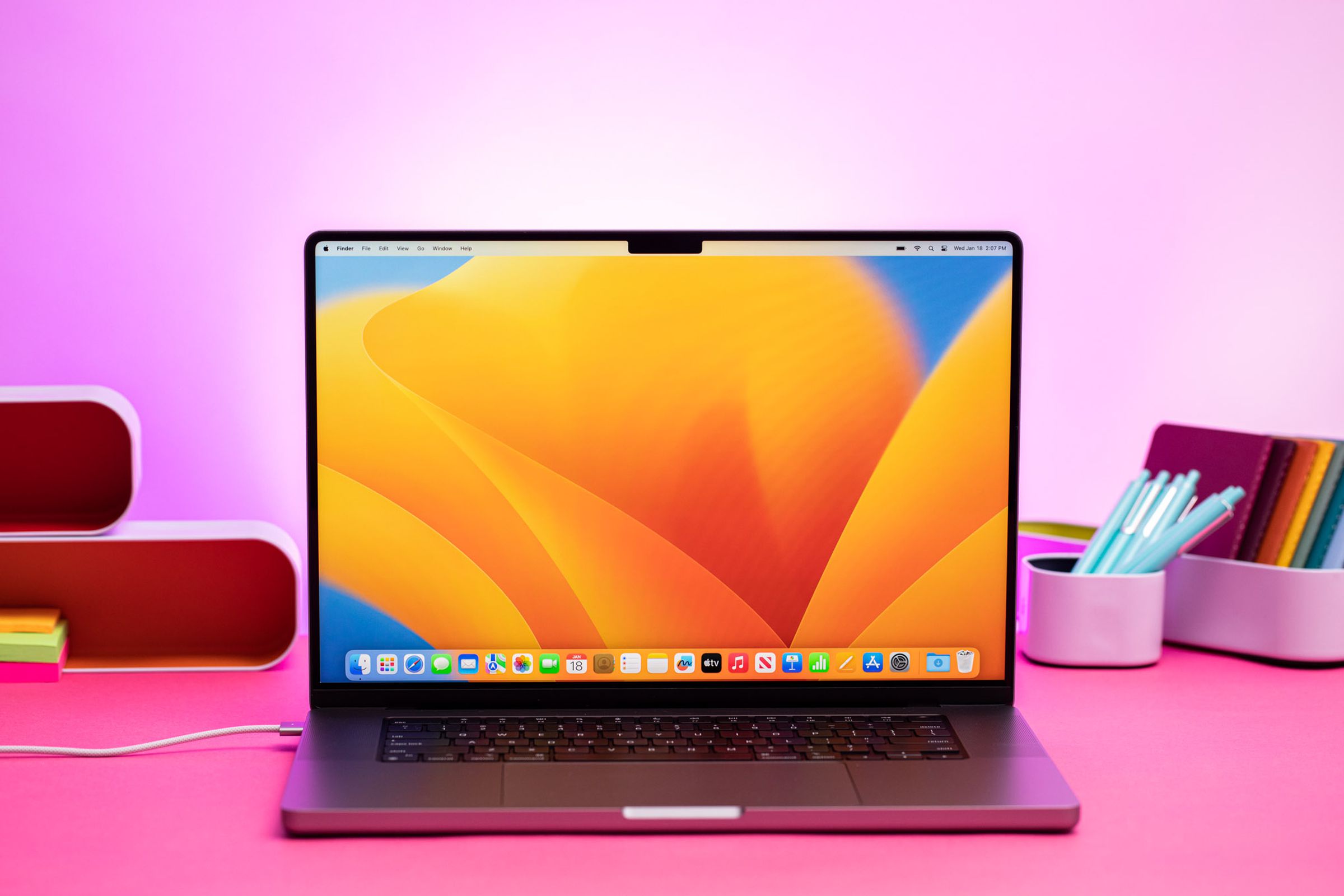 The MacBook Pro 16 (2023) on a pink table. The screen displays a blue and yellow desktop pattern.
